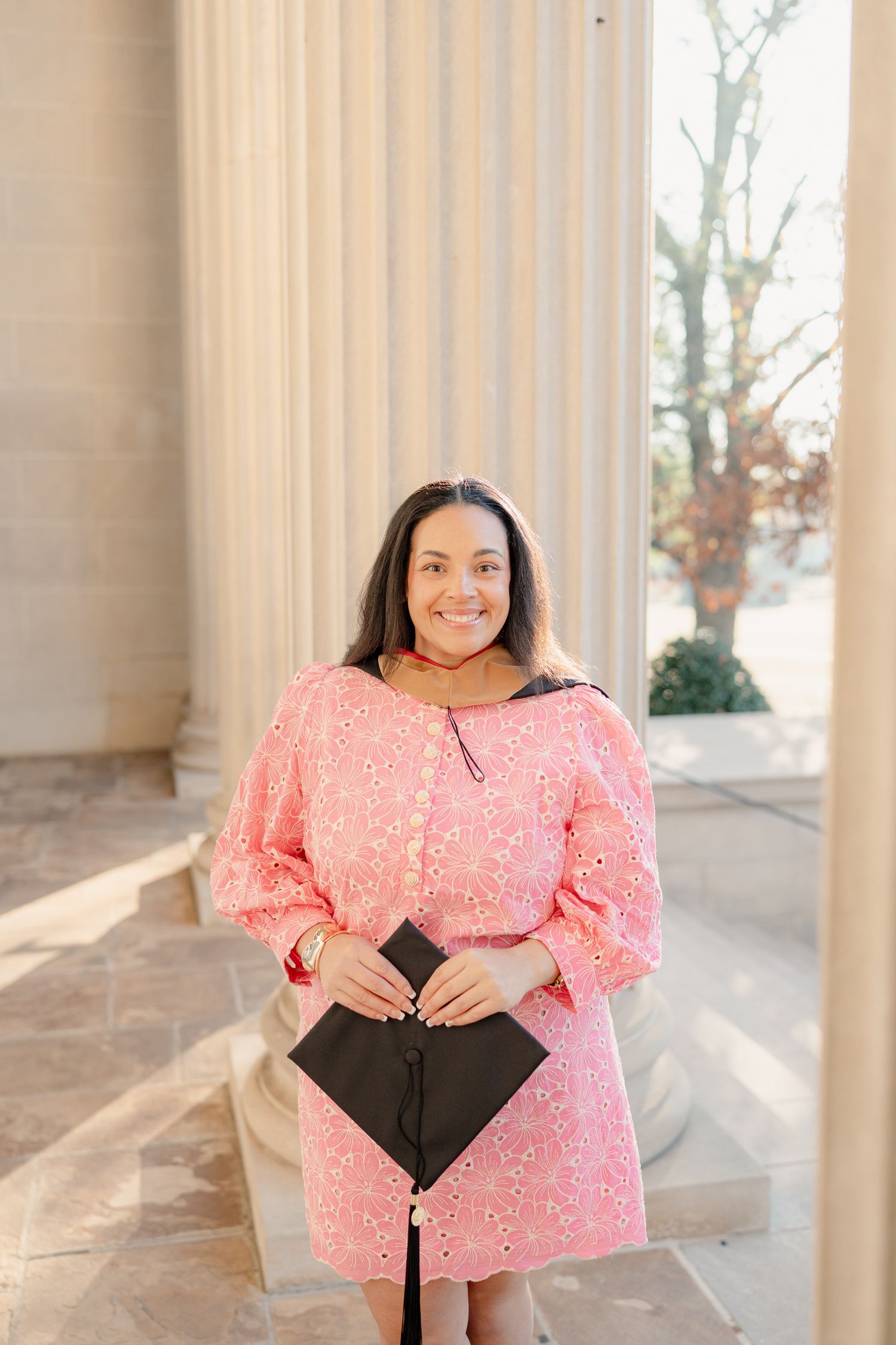 A woman in a pink eyelet dress poses outdoors near white columns on a sunny day.