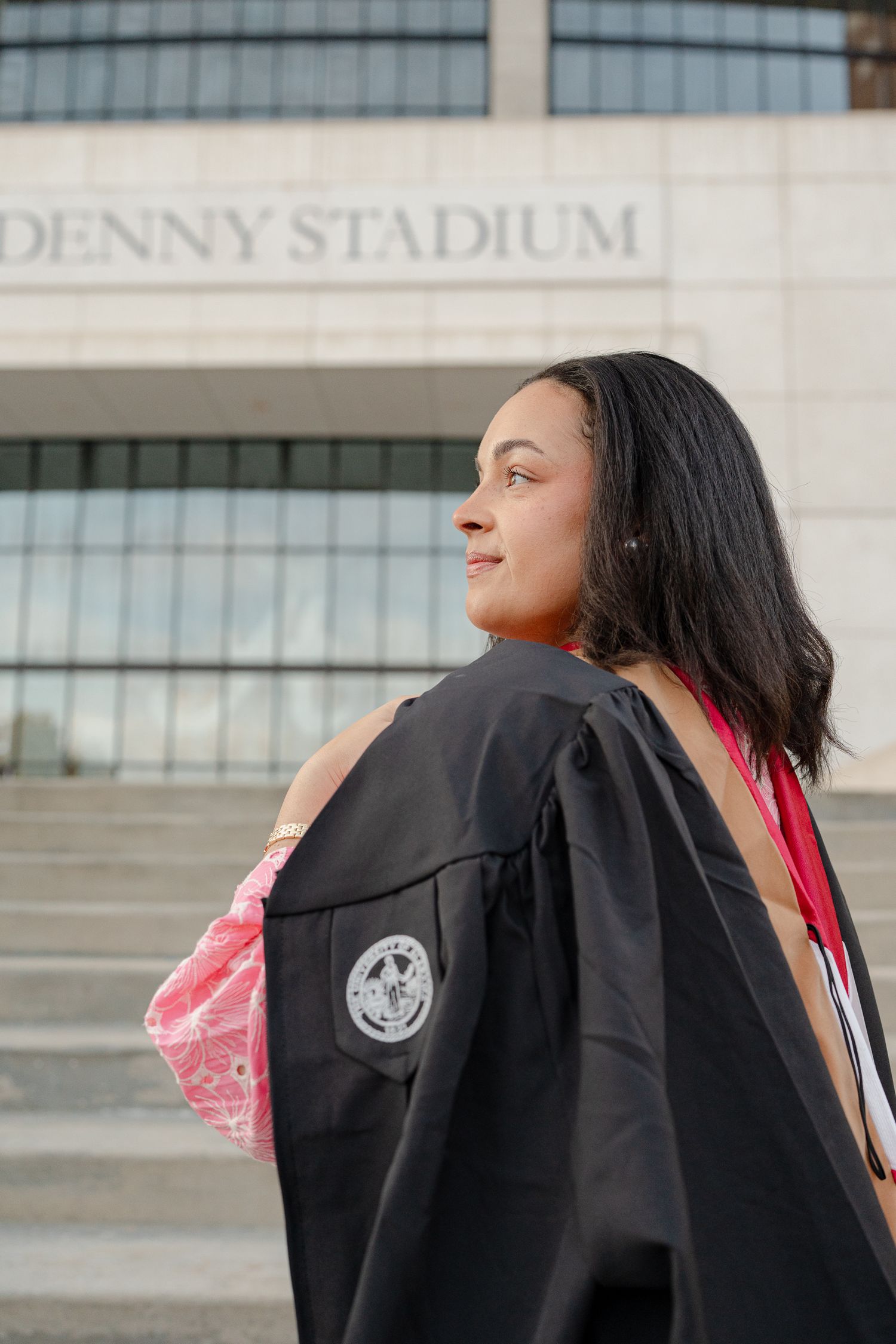 Graduate in black cap and gown standing outside Denny Stadium looking upward with joyful expression.