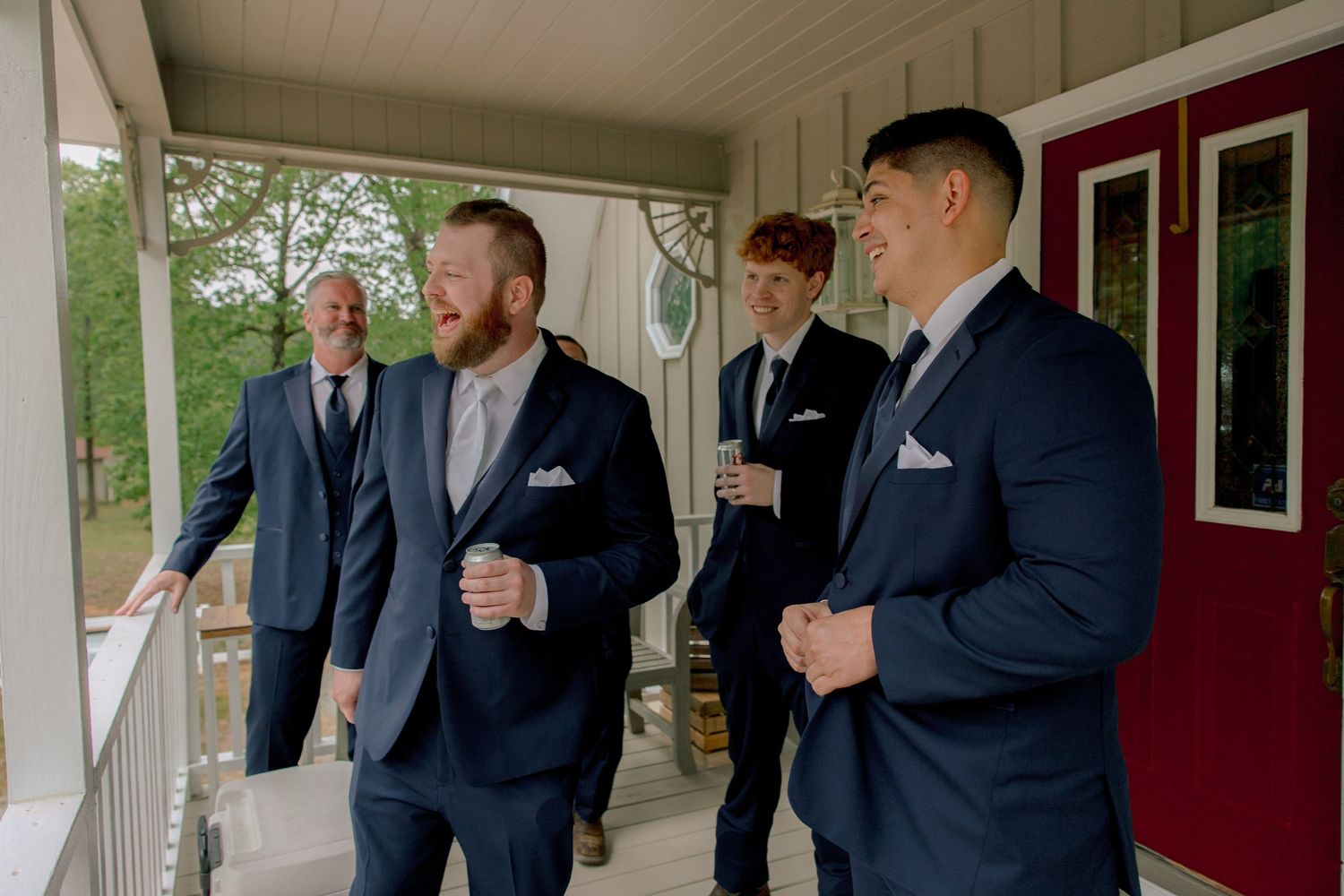 Four groomsmen in navy blue suits laughing and talking on a white porch before a wedding ceremony.