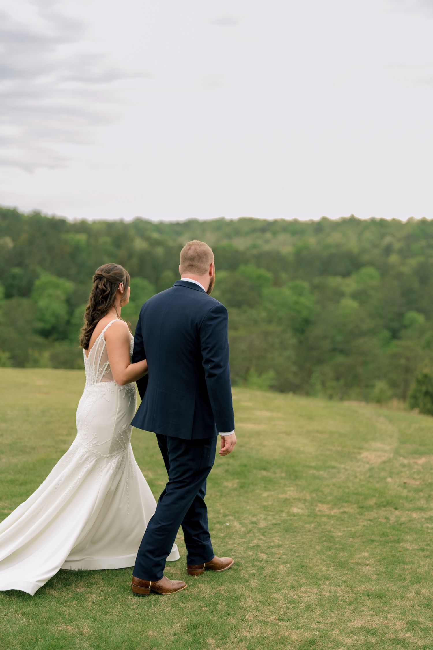 Bride and groom walking hand in hand across lush green hillside with wooded backdrop on wedding day.