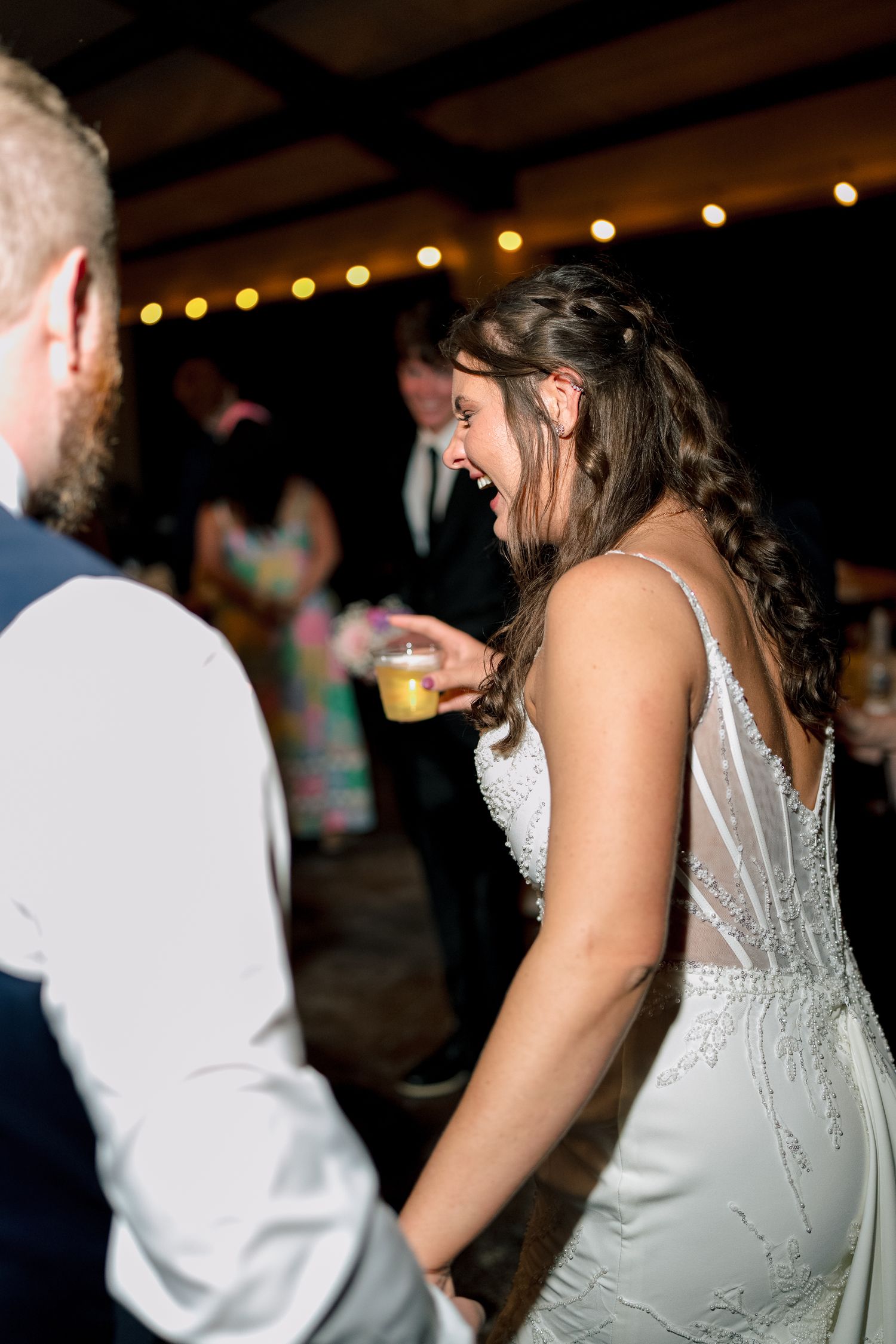 Bride laughs joyfully while dancing with groom at wedding reception under warm string lights.