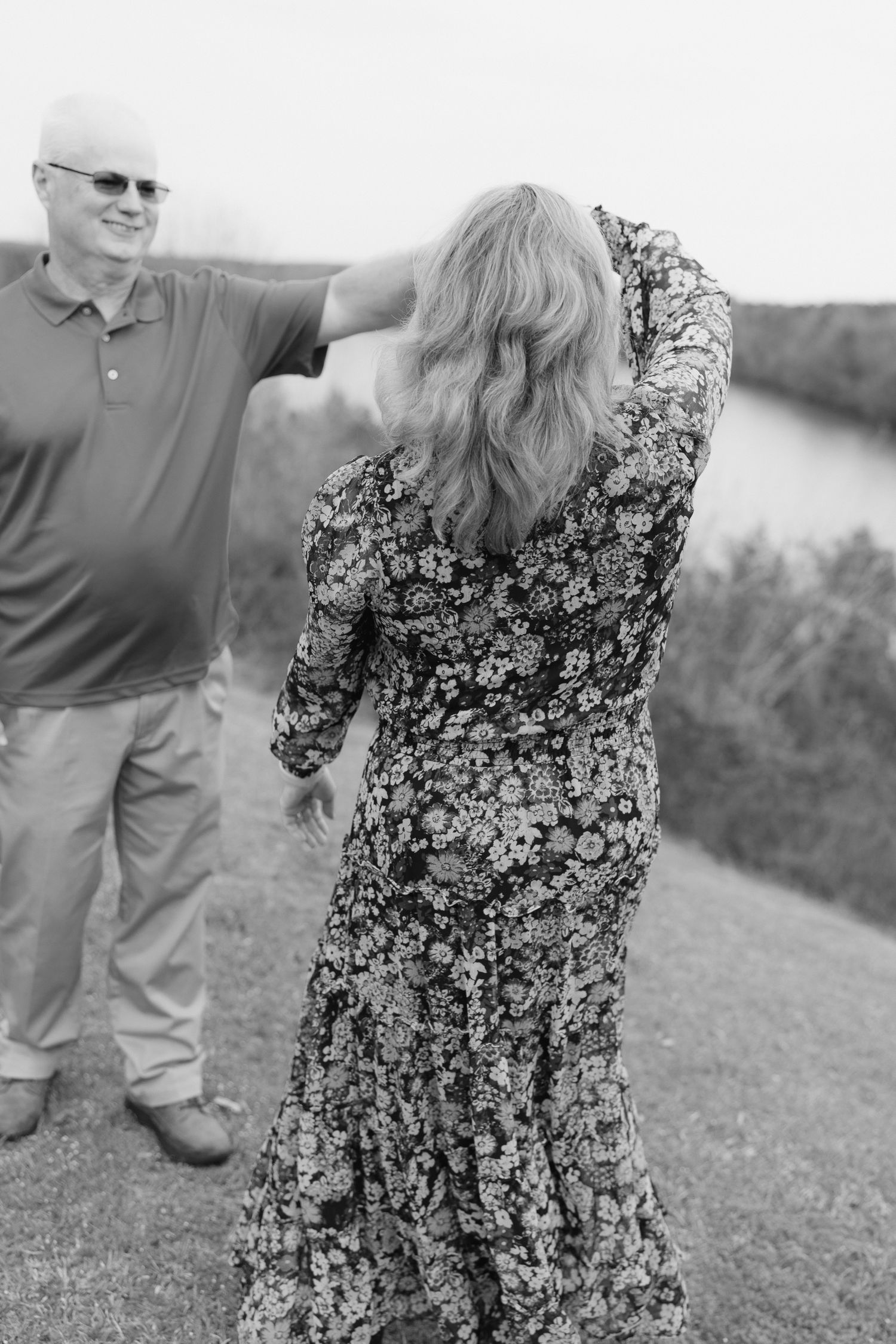 Black and white photo of couple near a river, woman in floral dress with hair blowing in the wind.