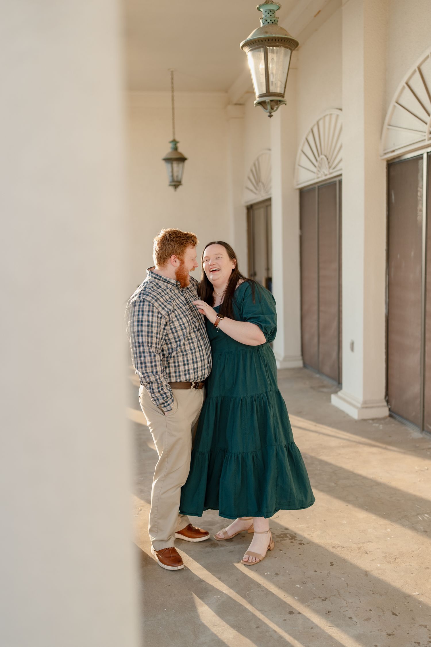 Couple laughing together on sunny columned porch, woman in teal maxi dress and man in plaid shirt sharing a joyful moment.