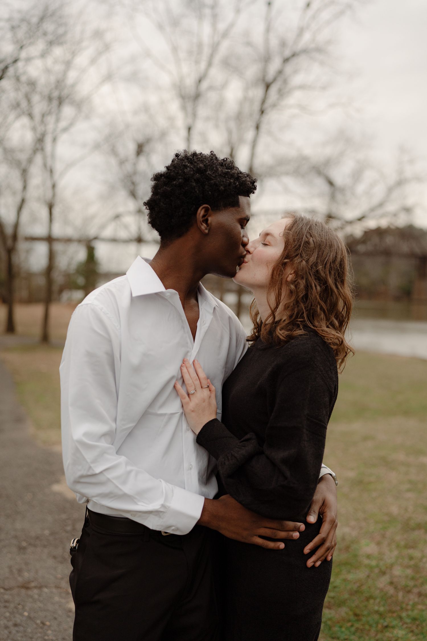 An interracial couple shares a tender kiss outdoors near bare winter trees along a riverside path.