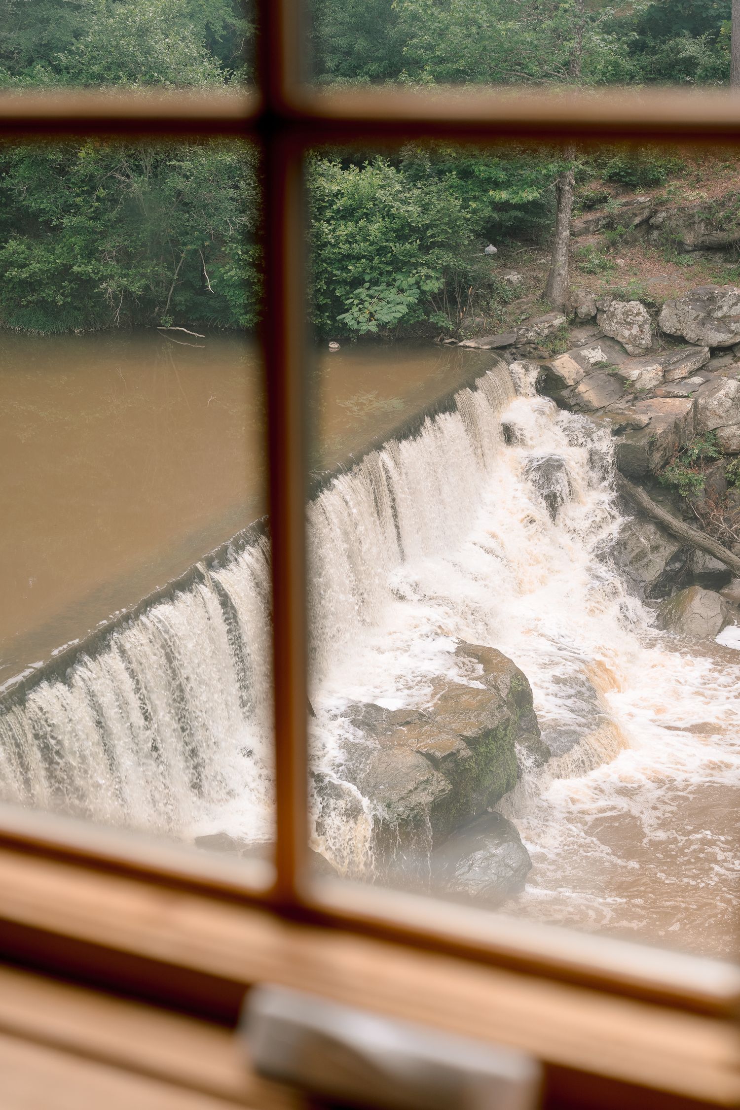 View through window of rushing waterfall cascading over rocks surrounded by lush green trees.