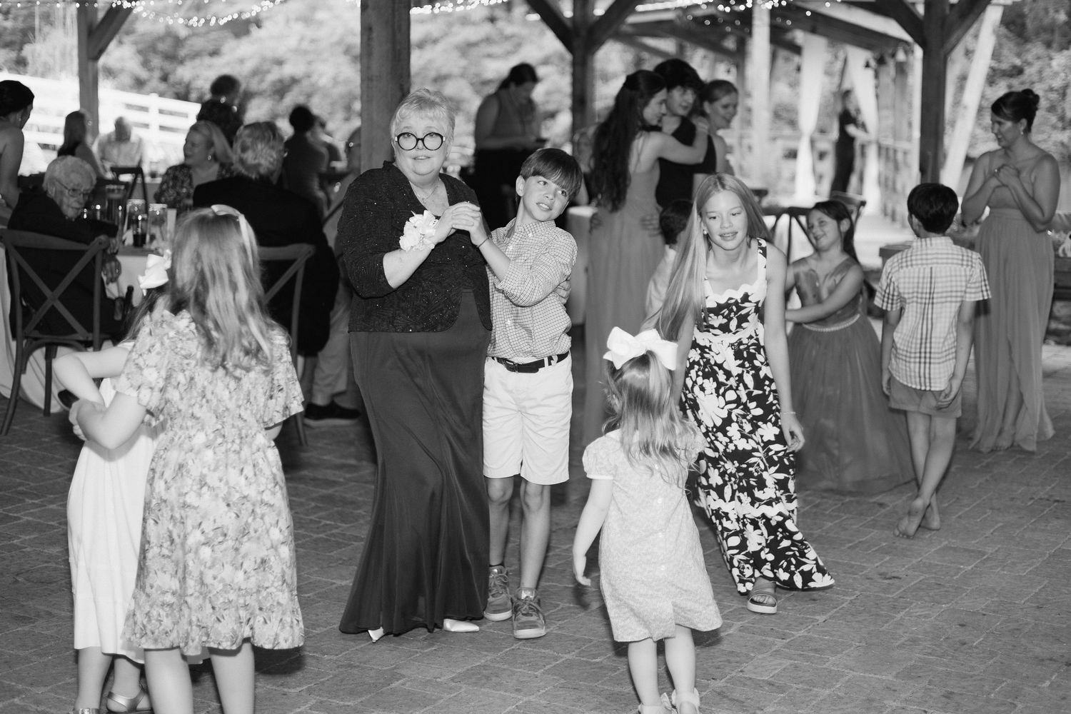Black and white photo of guests including children dancing joyfully at outdoor wedding reception.