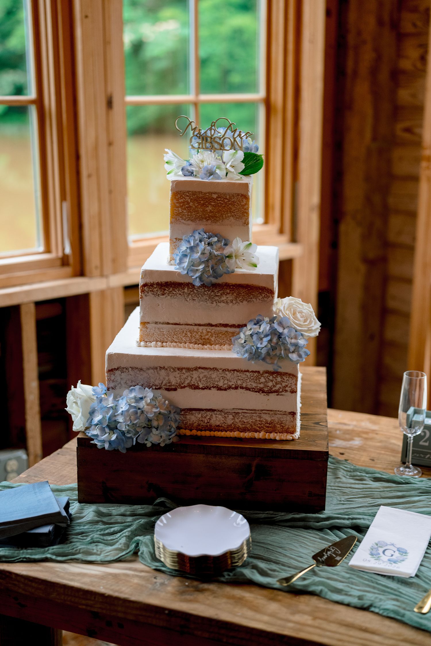 Three-tier square naked wedding cake decorated with blue hydrangeas and white roses on wooden stand.