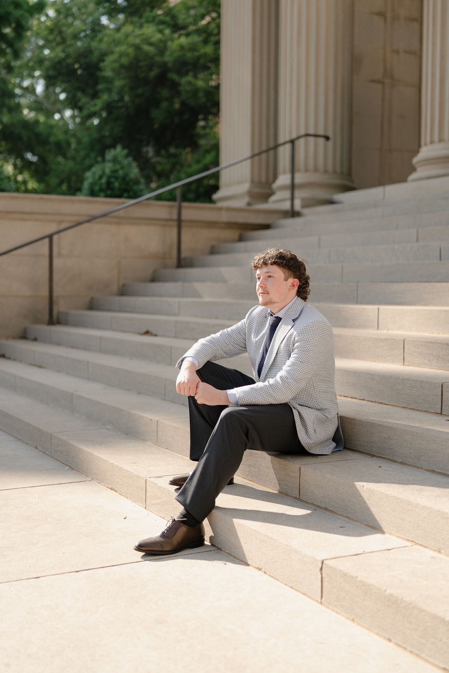 Young man in gray blazer and tie sitting thoughtfully on stone steps of a classical building with columns.