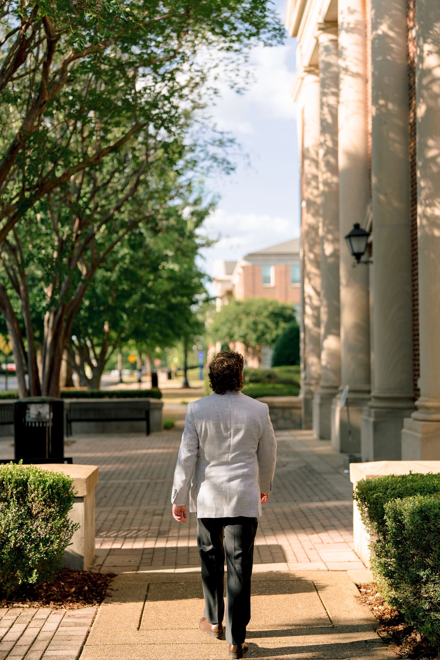 Graduate in grey blazer walks away from camera along brick pathway flanked by white columns.