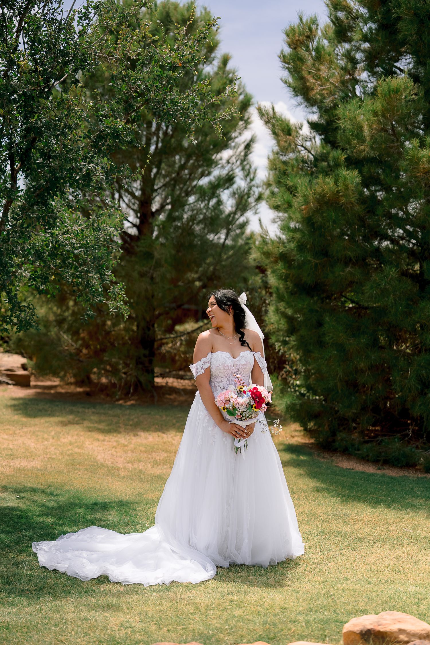 Bride in off-shoulder white ball gown holding floral bouquet standing on green lawn surrounded by pine trees.