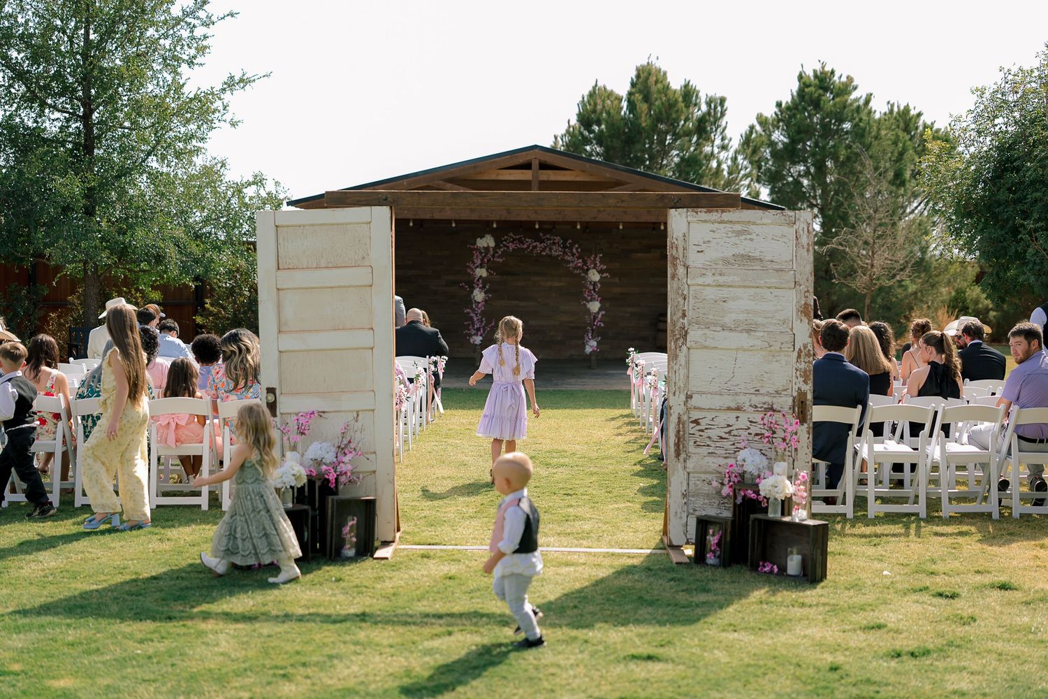 Outdoor wedding ceremony with white doors, floral decor, children walking down aisle, guests seated on white chairs, rustic barn backdrop.