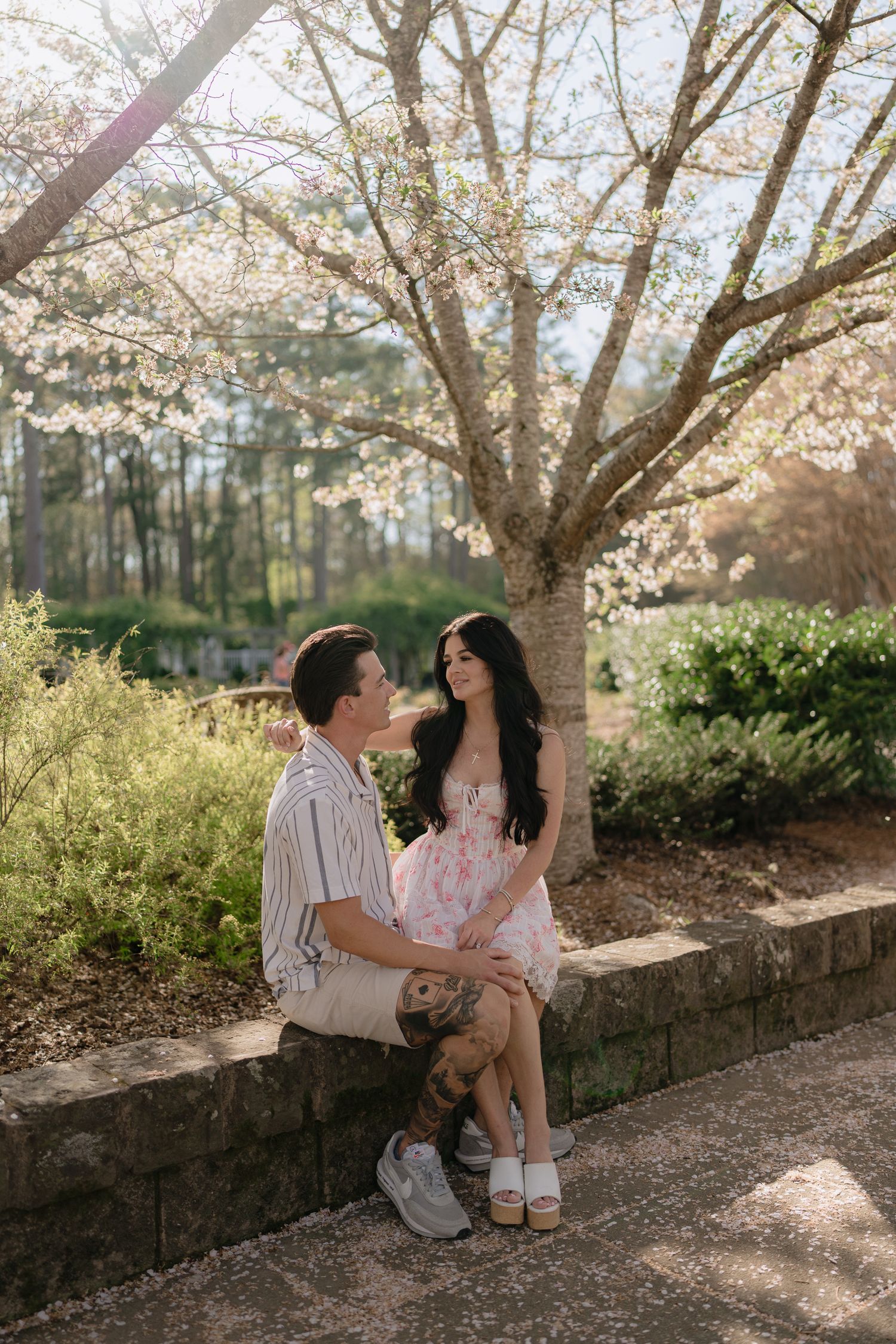Couple sitting on stone wall under blooming spring tree, woman in floral dress, man in striped shirt, golden hour light.
