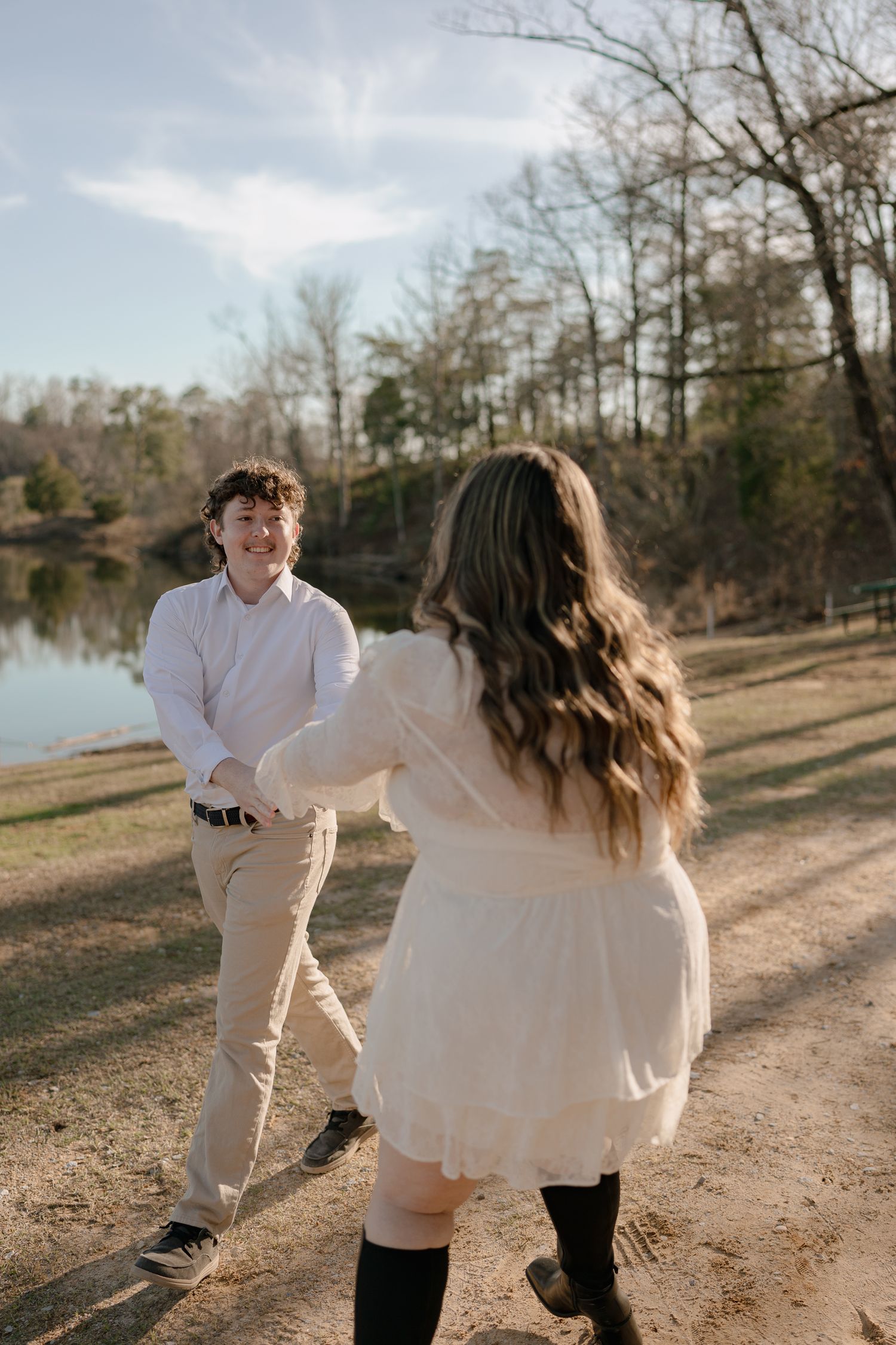 Couple holding hands near a lake, woman in white dress and knee-high boots, man in white shirt and khakis.
