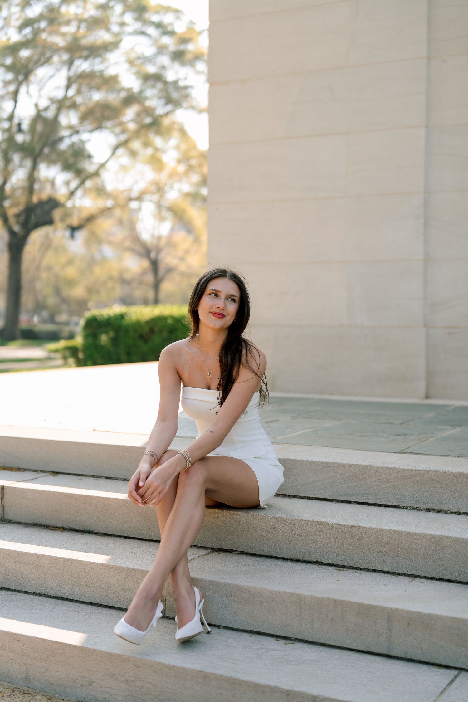 A woman in a white strapless dress sits elegantly on stone steps in warm afternoon sunlight.