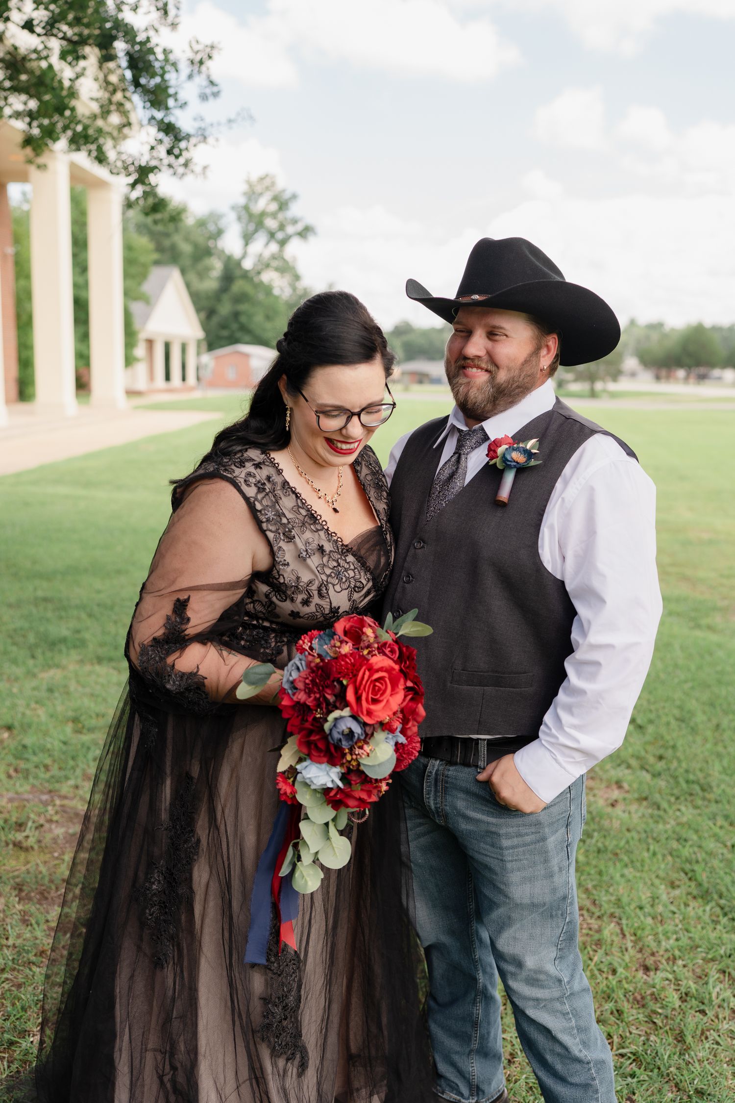 Couple in western-themed wedding attire share romantic moment with red floral bouquet outdoors.