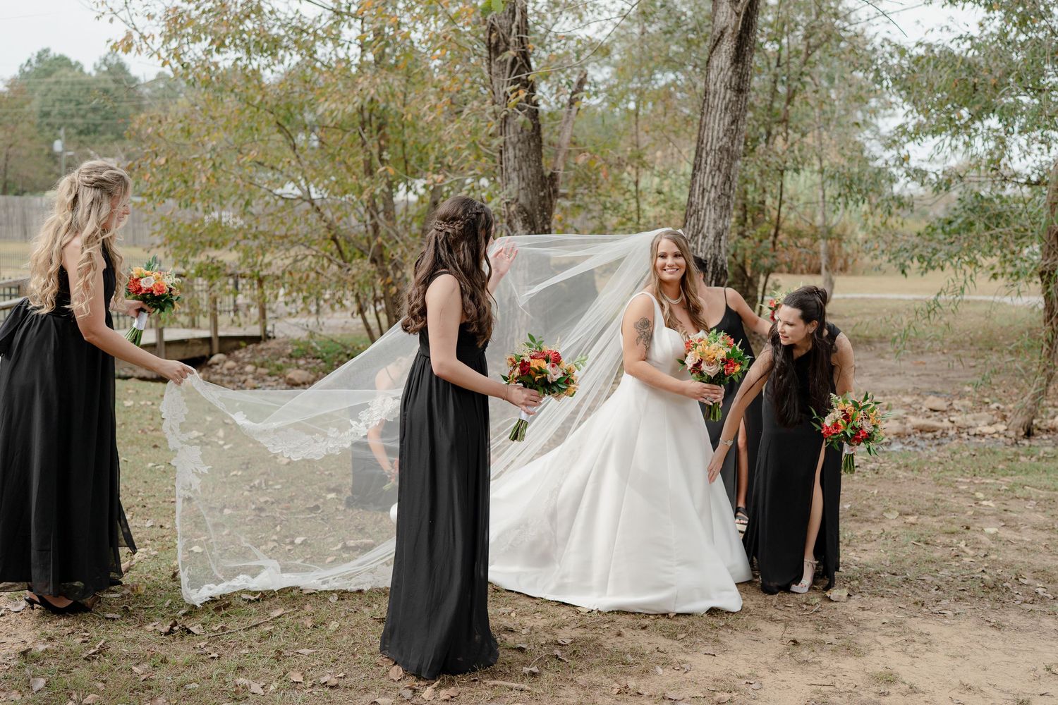 Wedding party in black dresses helps adjust the bride's veil in an outdoor woodland setting.
