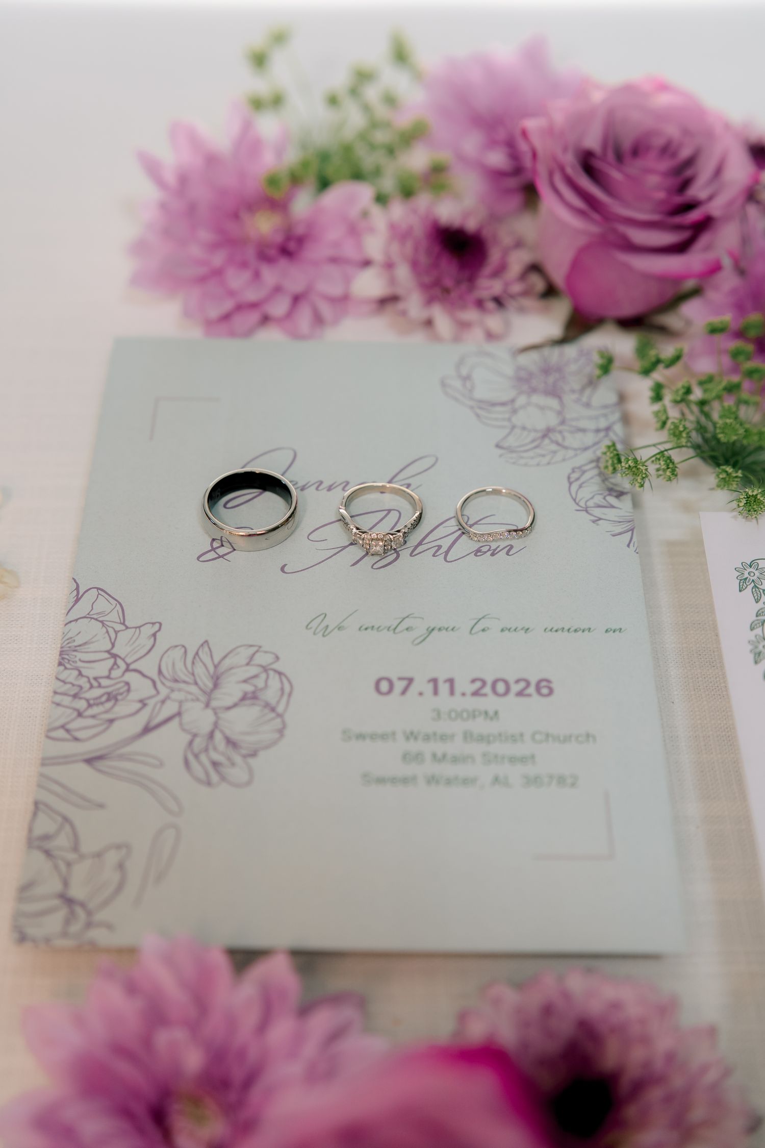 Wedding rings rest on a floral invitation for a July 2026 ceremony, surrounded by purple roses and pink chrysanthemums.