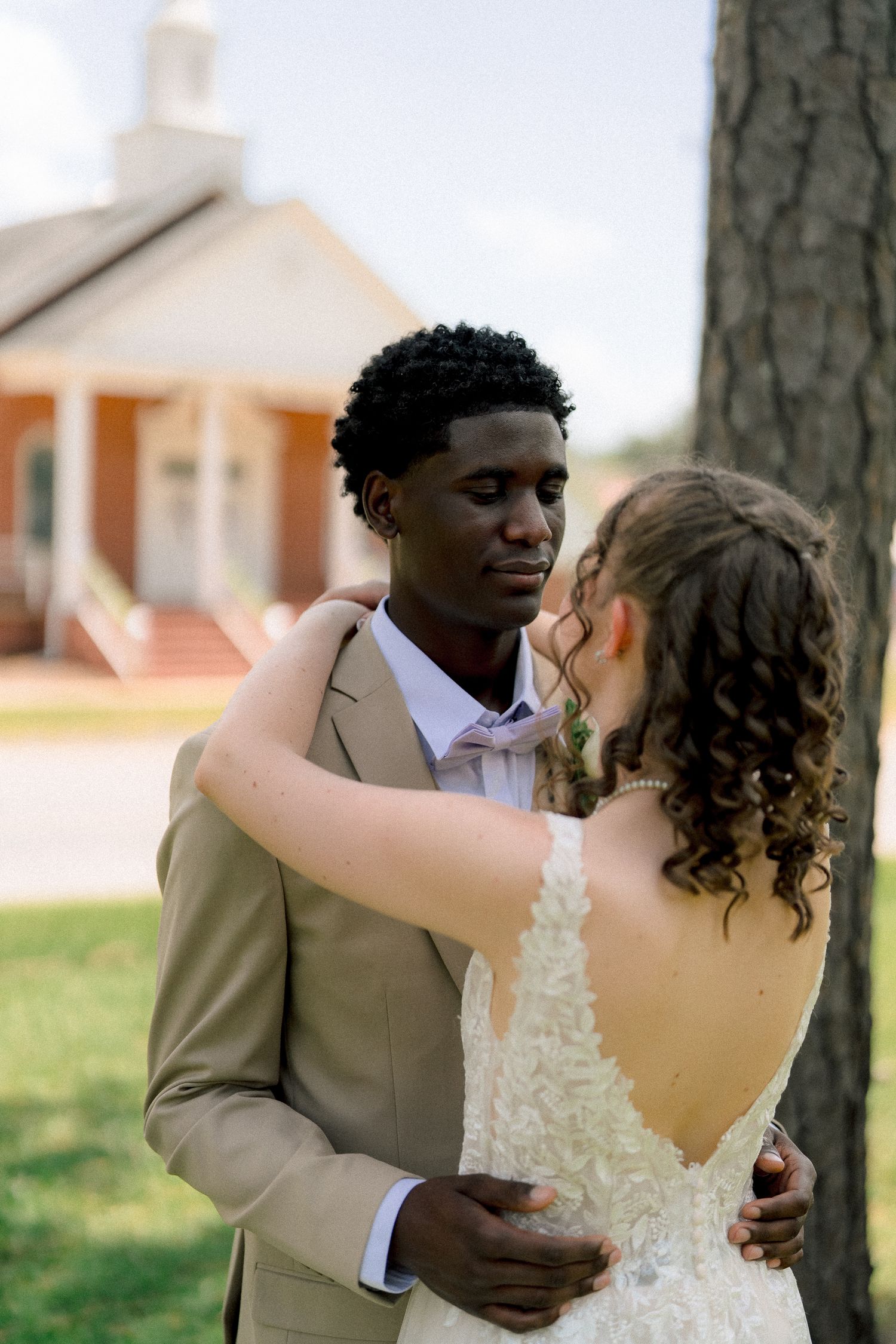 Groom in tan suit and bride in lace backless gown embrace near tree outside brick church.