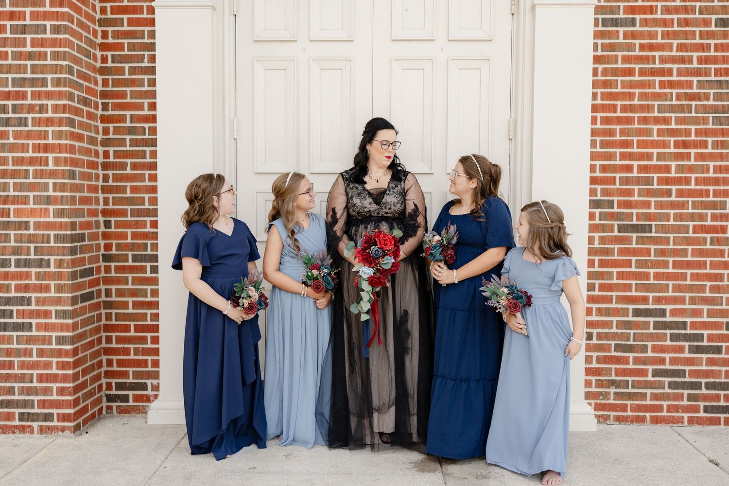 A wedding party in blue and gray dresses poses together against a brick wall with white doors.