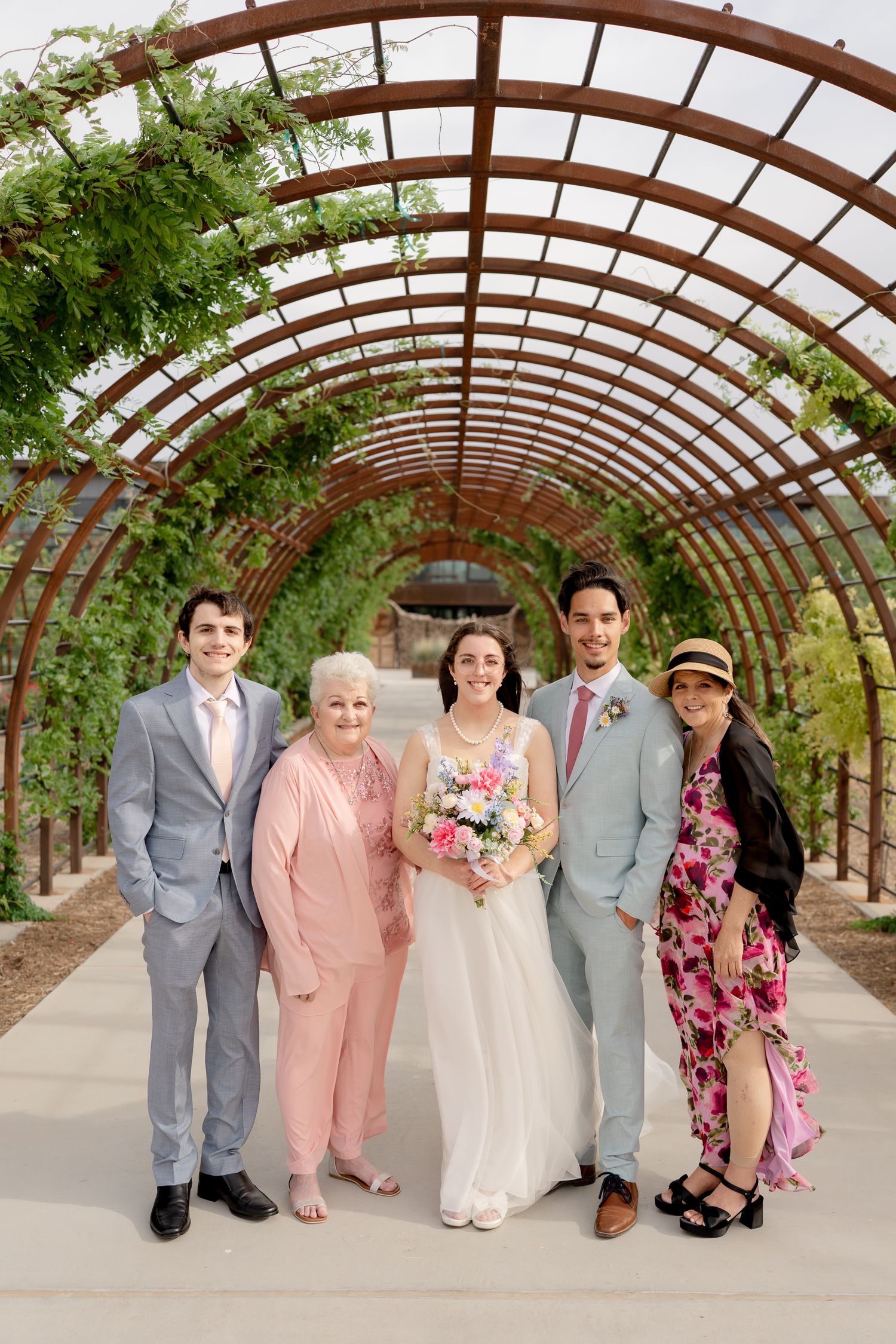 Wedding party poses under curved wooden pergola with greenery and flowers in soft pink and gray attire.