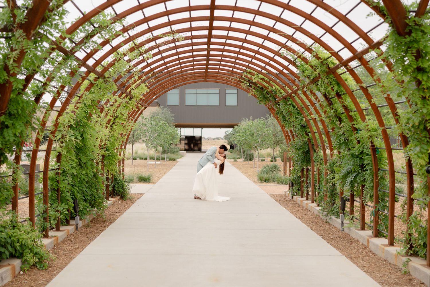 A curved wooden arbor walkway with climbing vines creates an elegant garden tunnel at an outdoor venue.