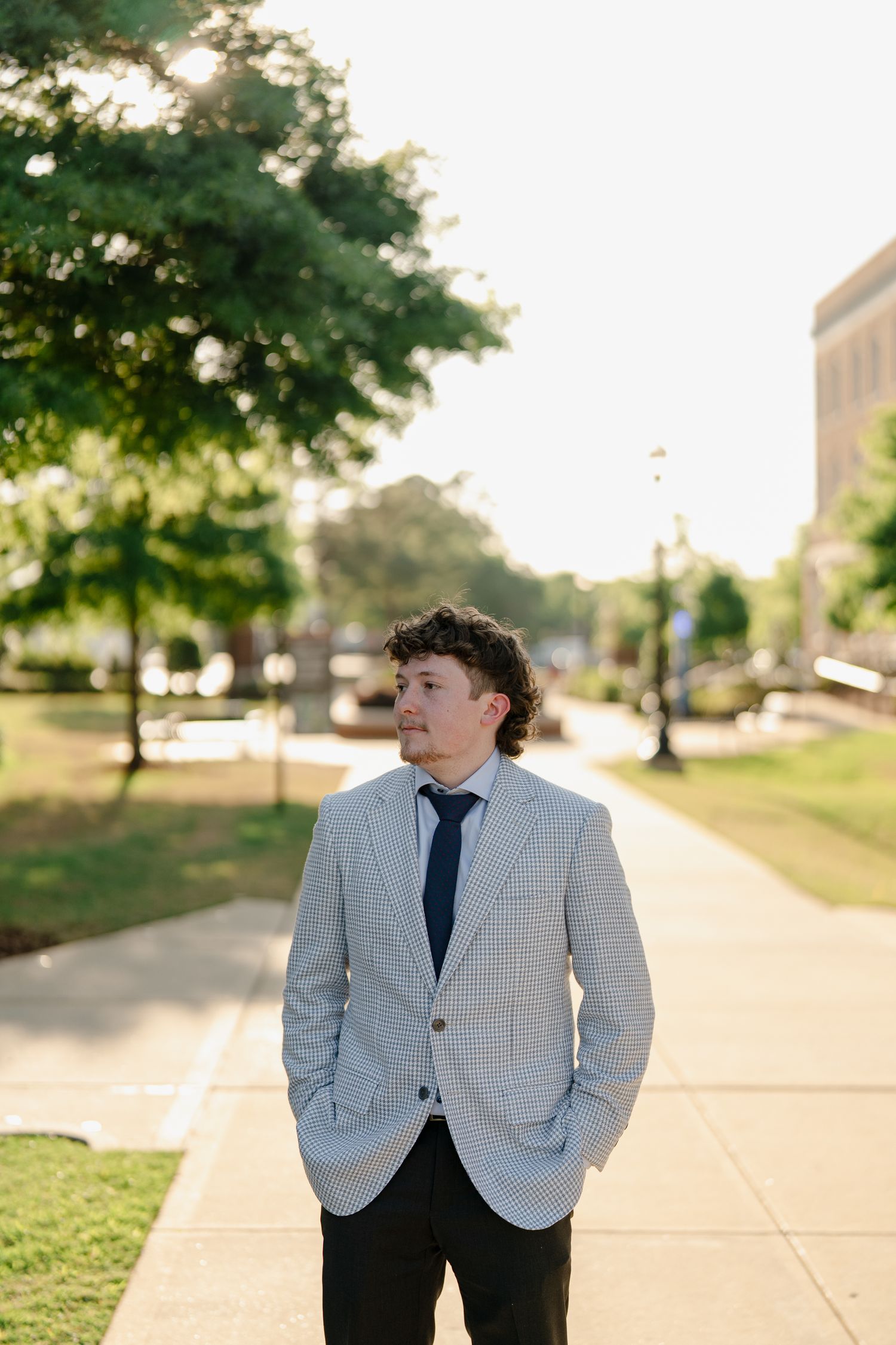 Male graduate in grey blazer and tie stands on sunlit campus walkway looking to the side.
