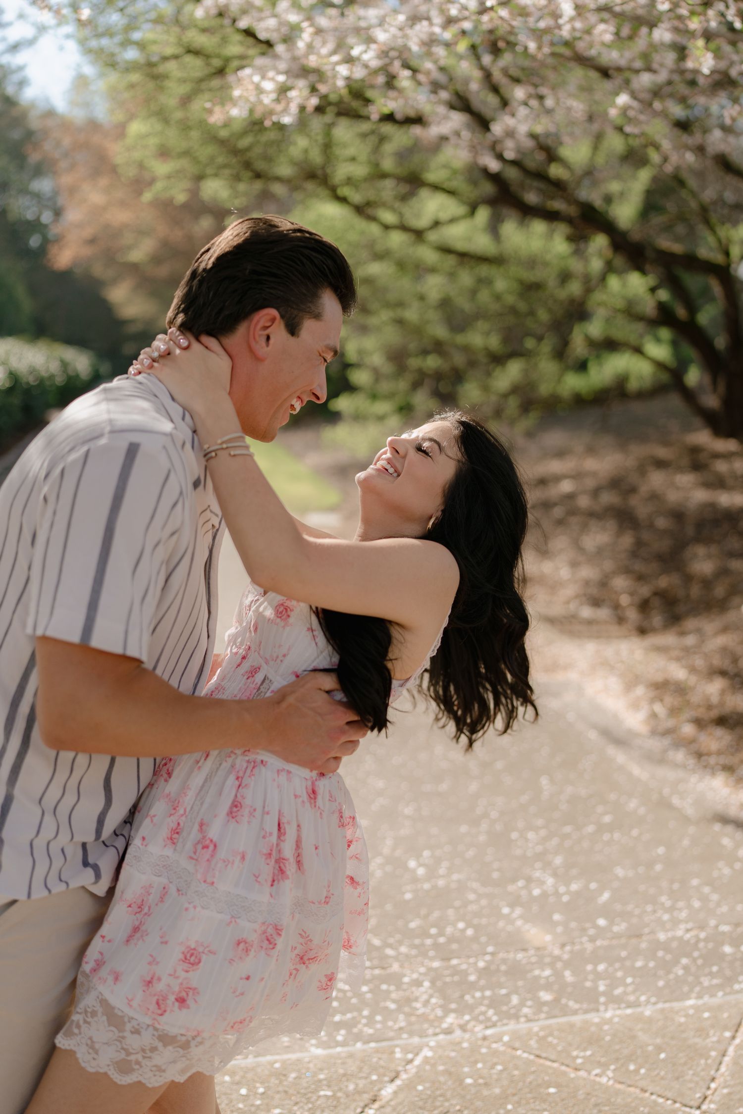 A joyful couple laughs and embraces on a sunlit path surrounded by spring cherry blossom trees.