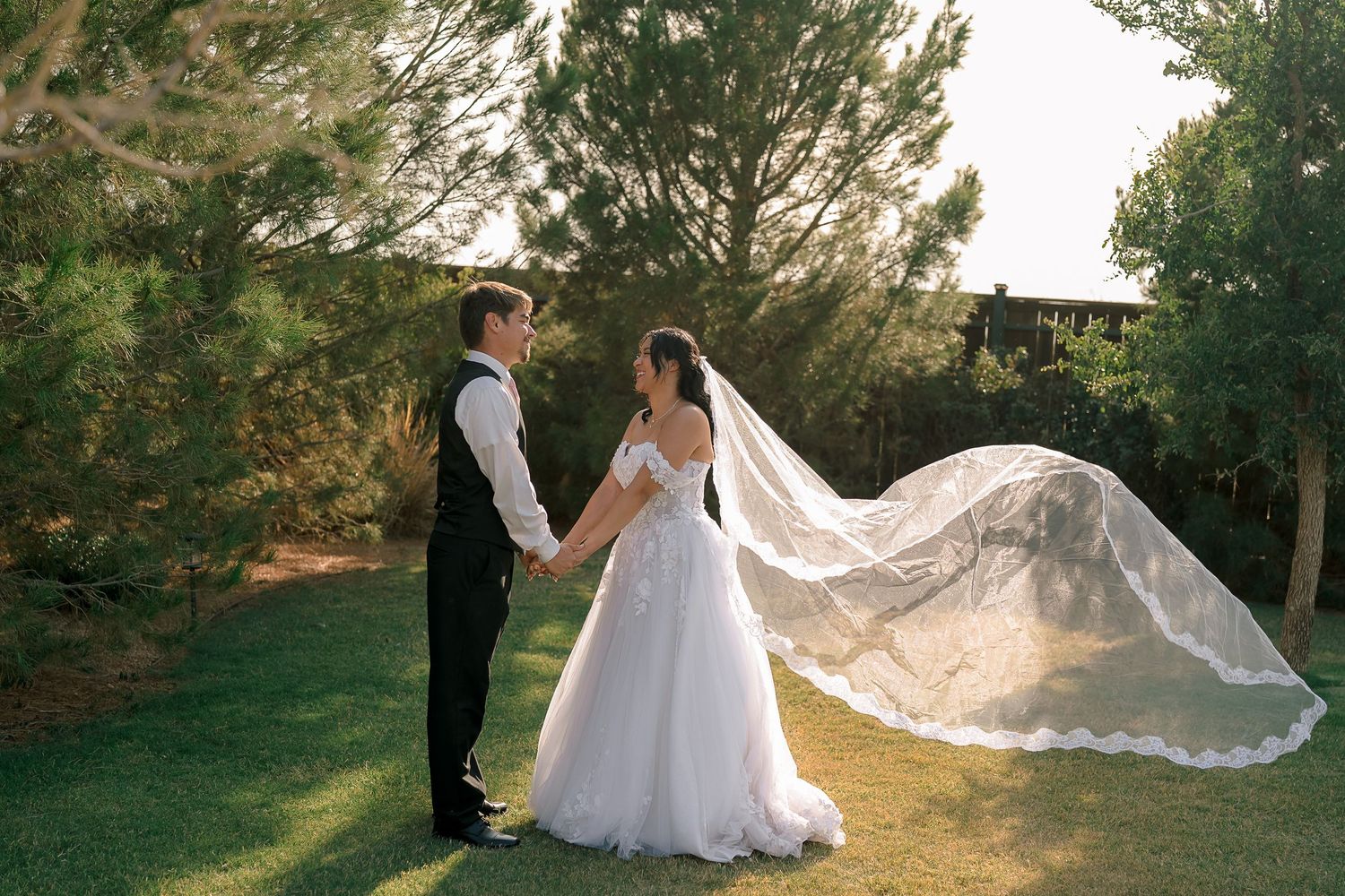Bride and groom hold hands outdoors as her lace cathedral veil billows in the wind among pine trees.
