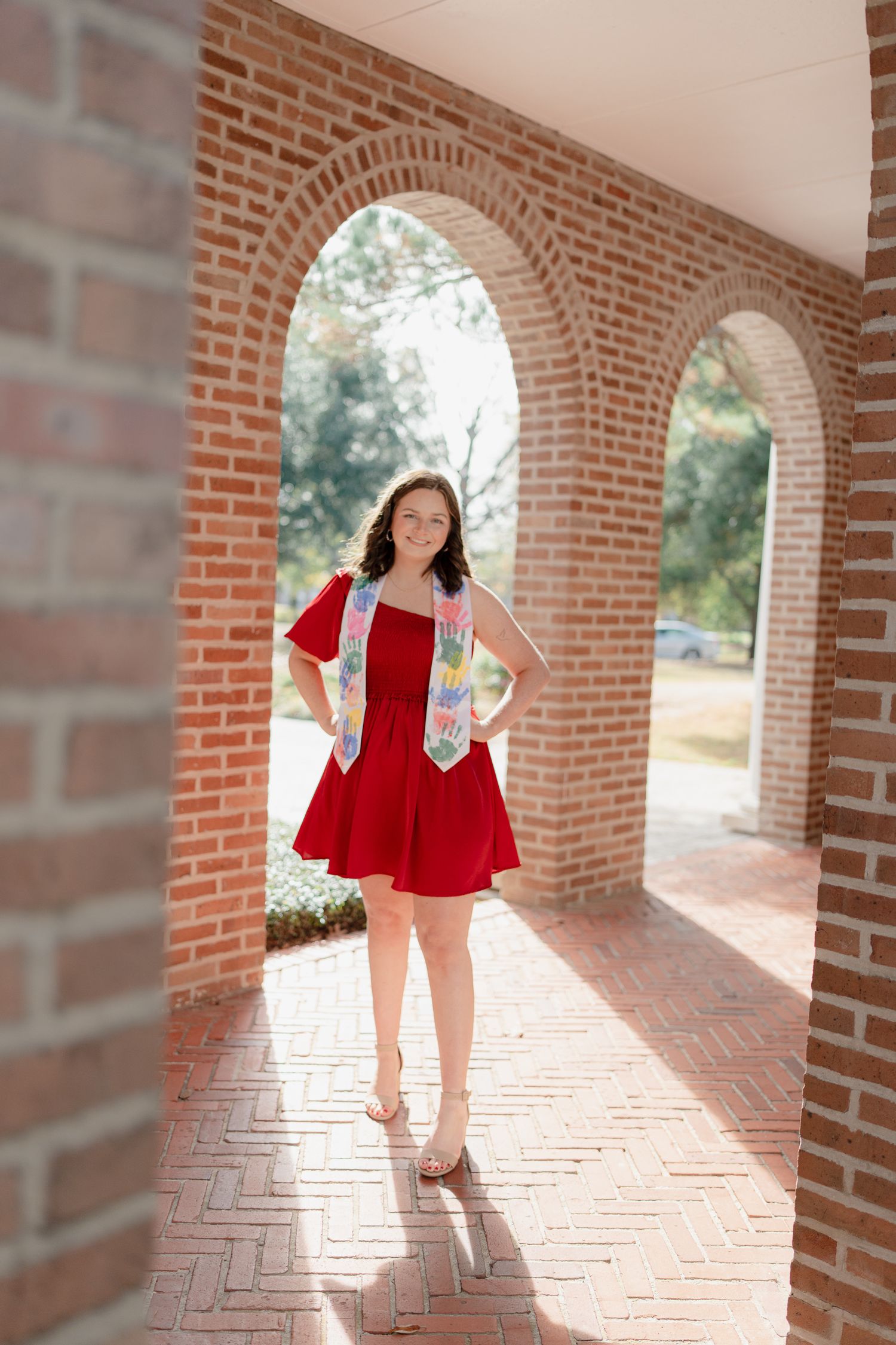 A person in a red dress and white cardigan poses playfully in a brick archway walkway on a sunny day.