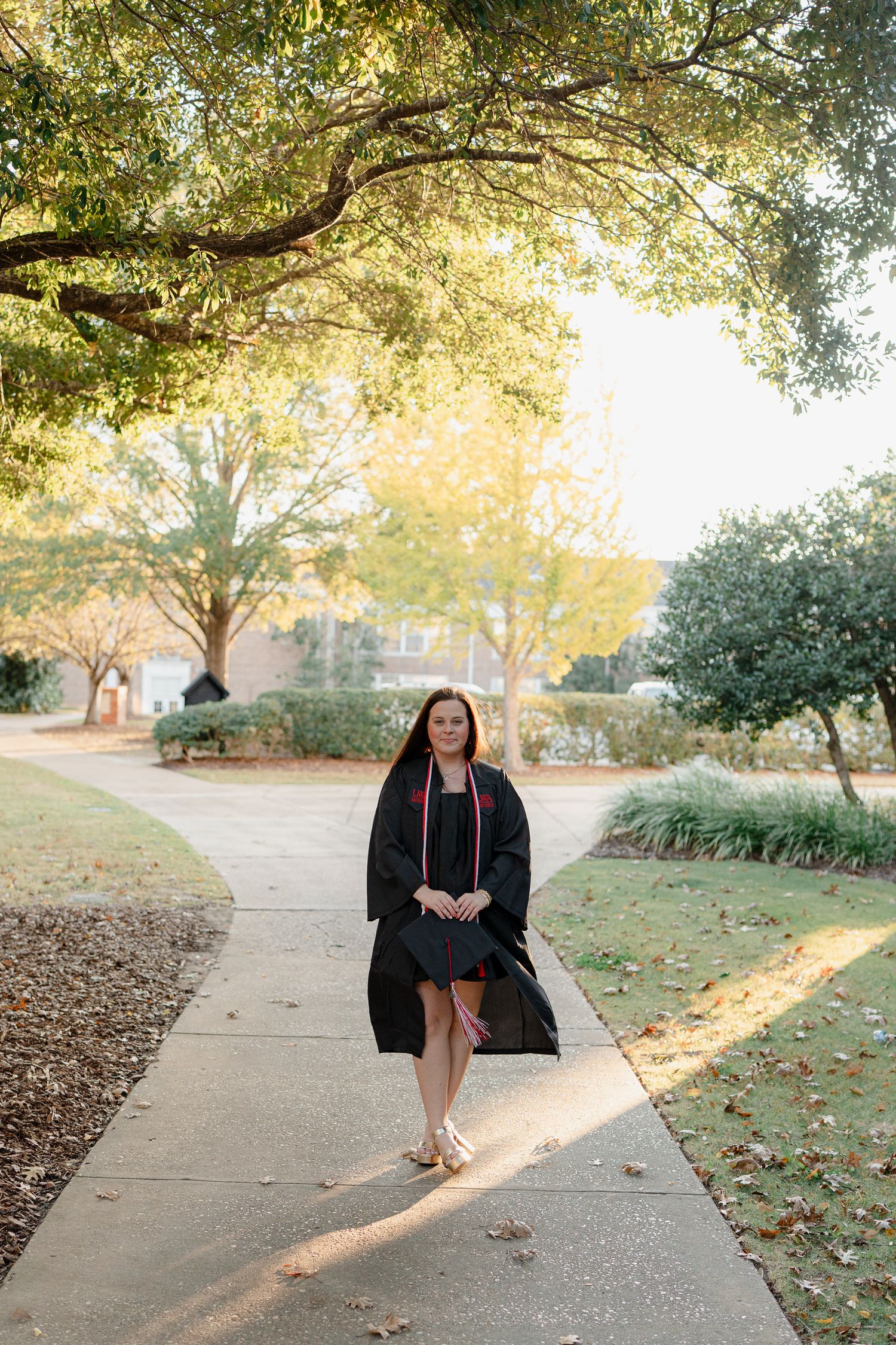 Graduate walks along tree-lined campus pathway during sunset graduation photoshoot.