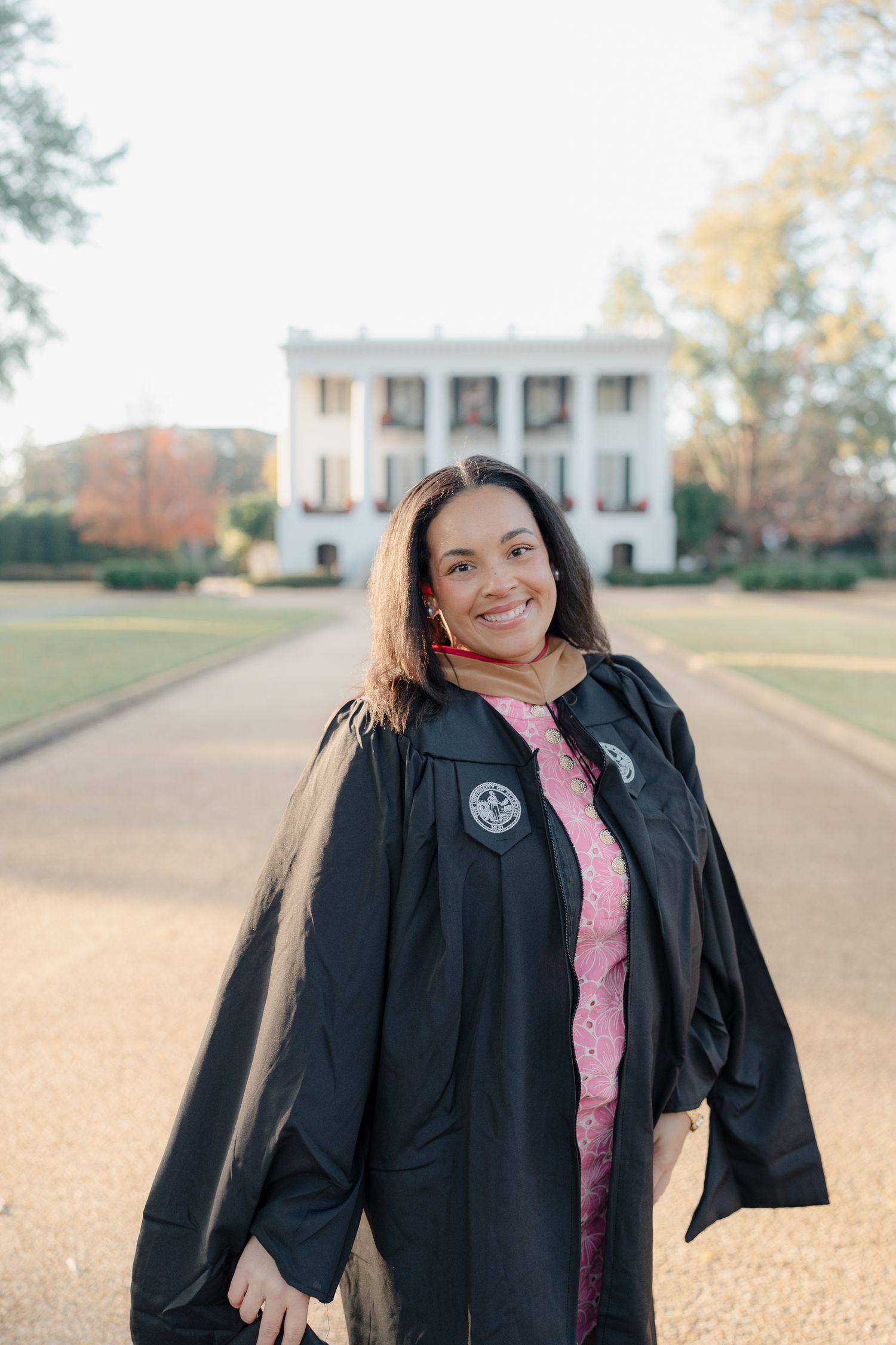Graduate in black academic regalia and pink dress smiles joyfully during outdoor graduation photoshoot on campus grounds.
