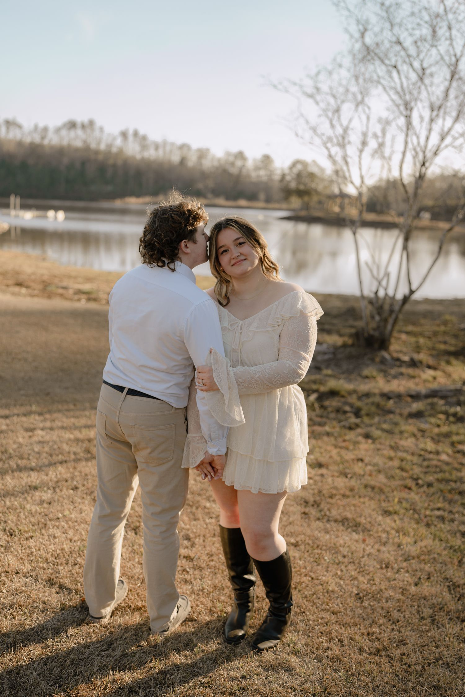 Smiling woman in white dress and black boots holds hands with man near a sunlit lake.