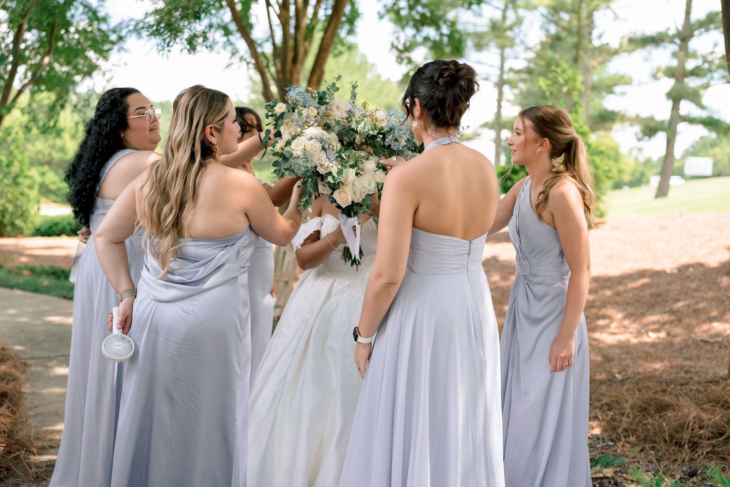 A group of bridesmaids in flowing light gray dresses laugh together during an outdoor wedding photo session.