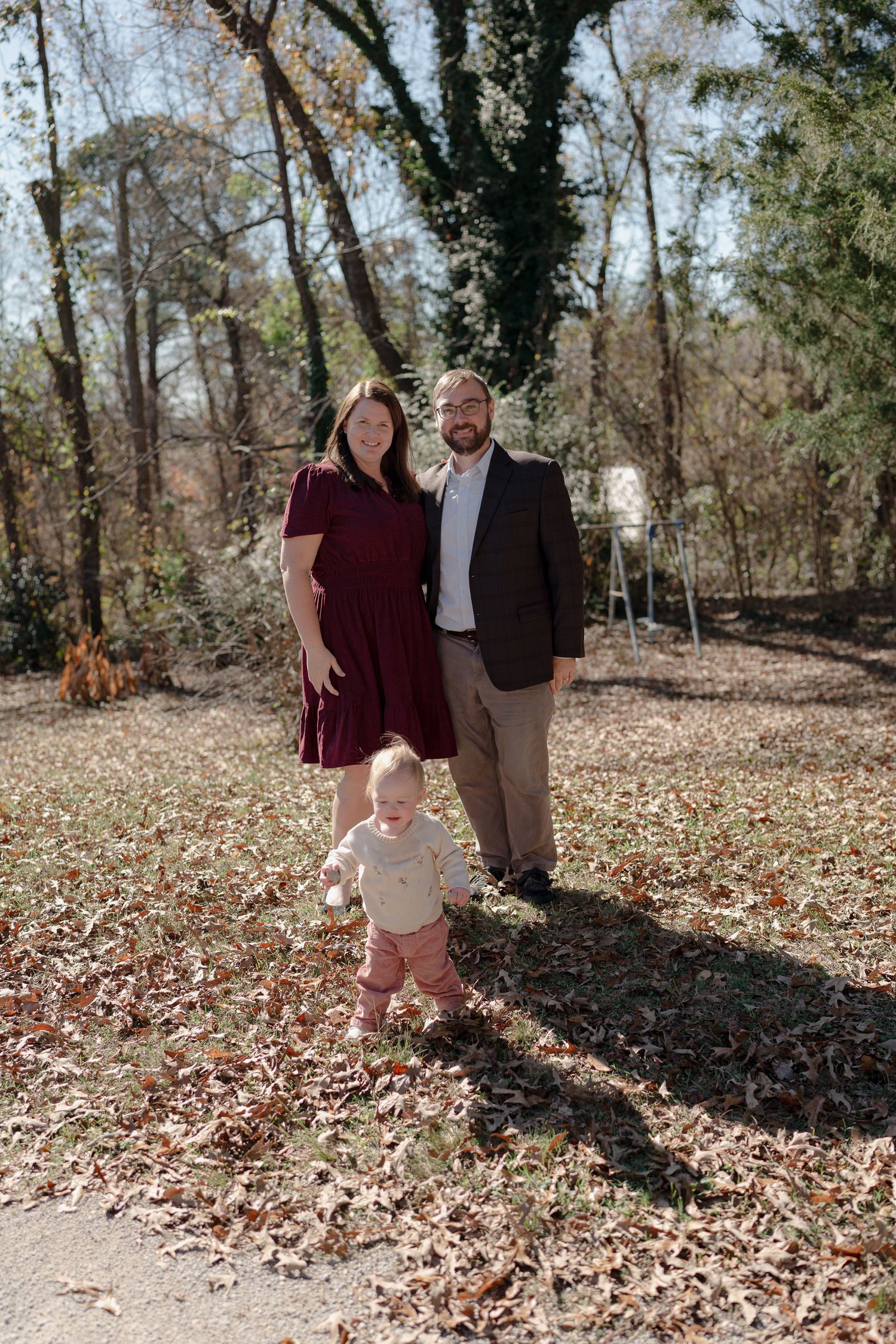 A family walks together through a wooded area covered in fallen autumn leaves on a sunny afternoon.