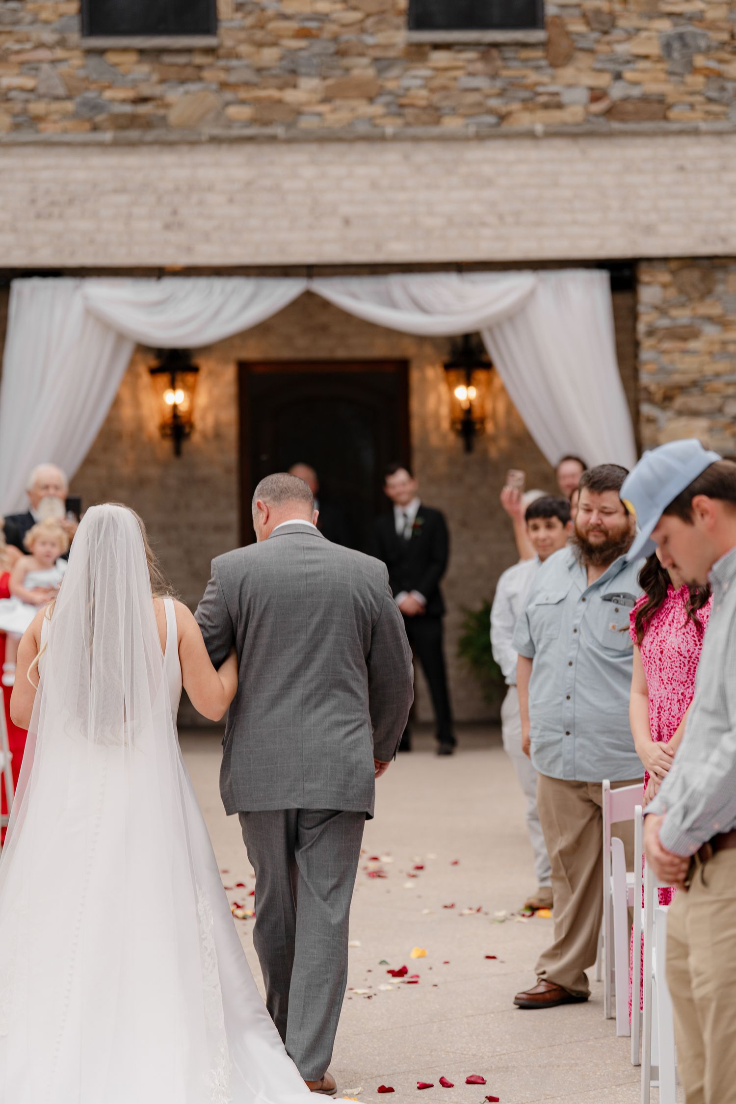 Wedding processional down an aisle decorated with rose petals at an elegant stone venue entrance.