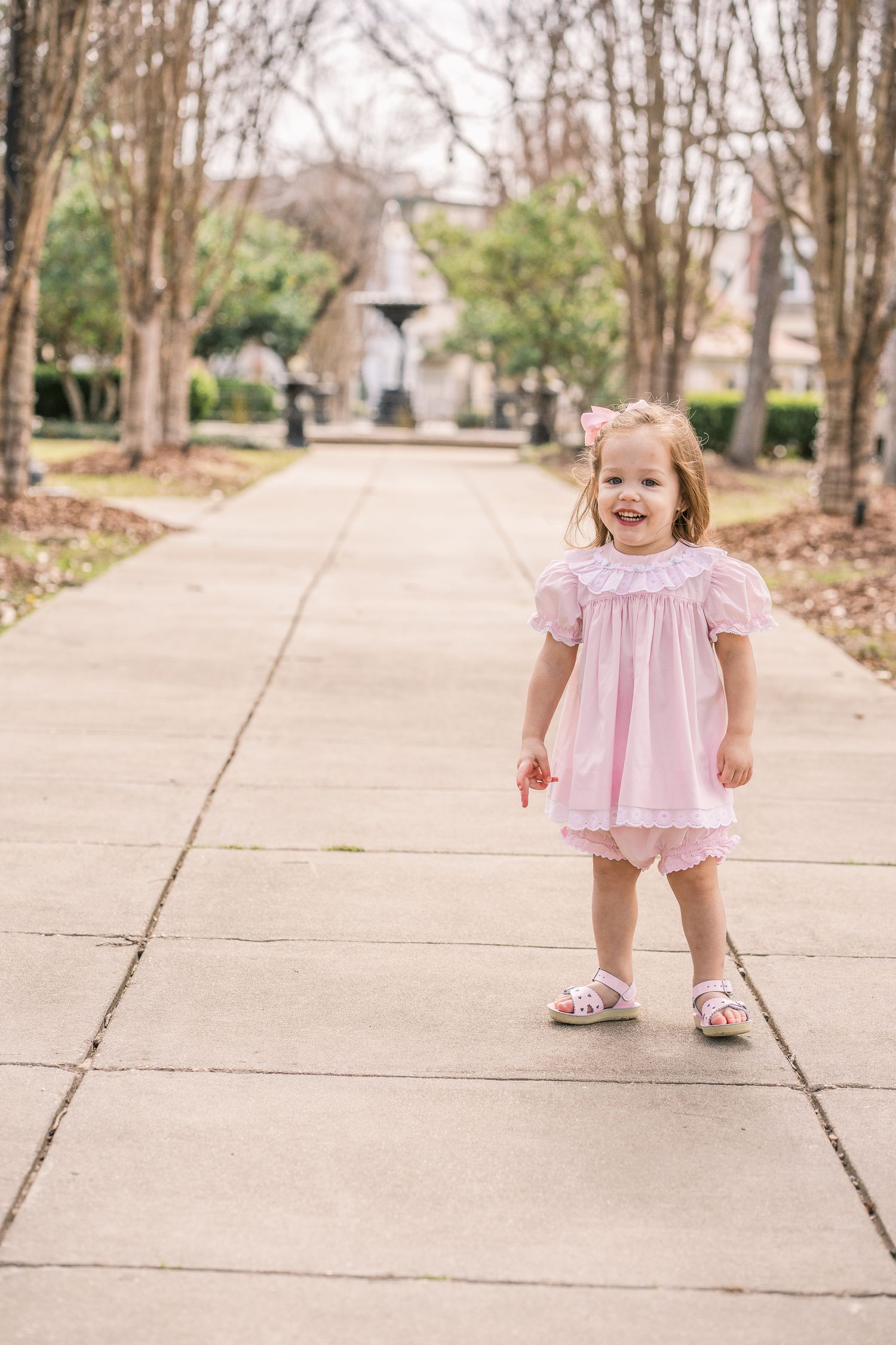 Child in pink dress smiles while standing on tree-lined walkway.