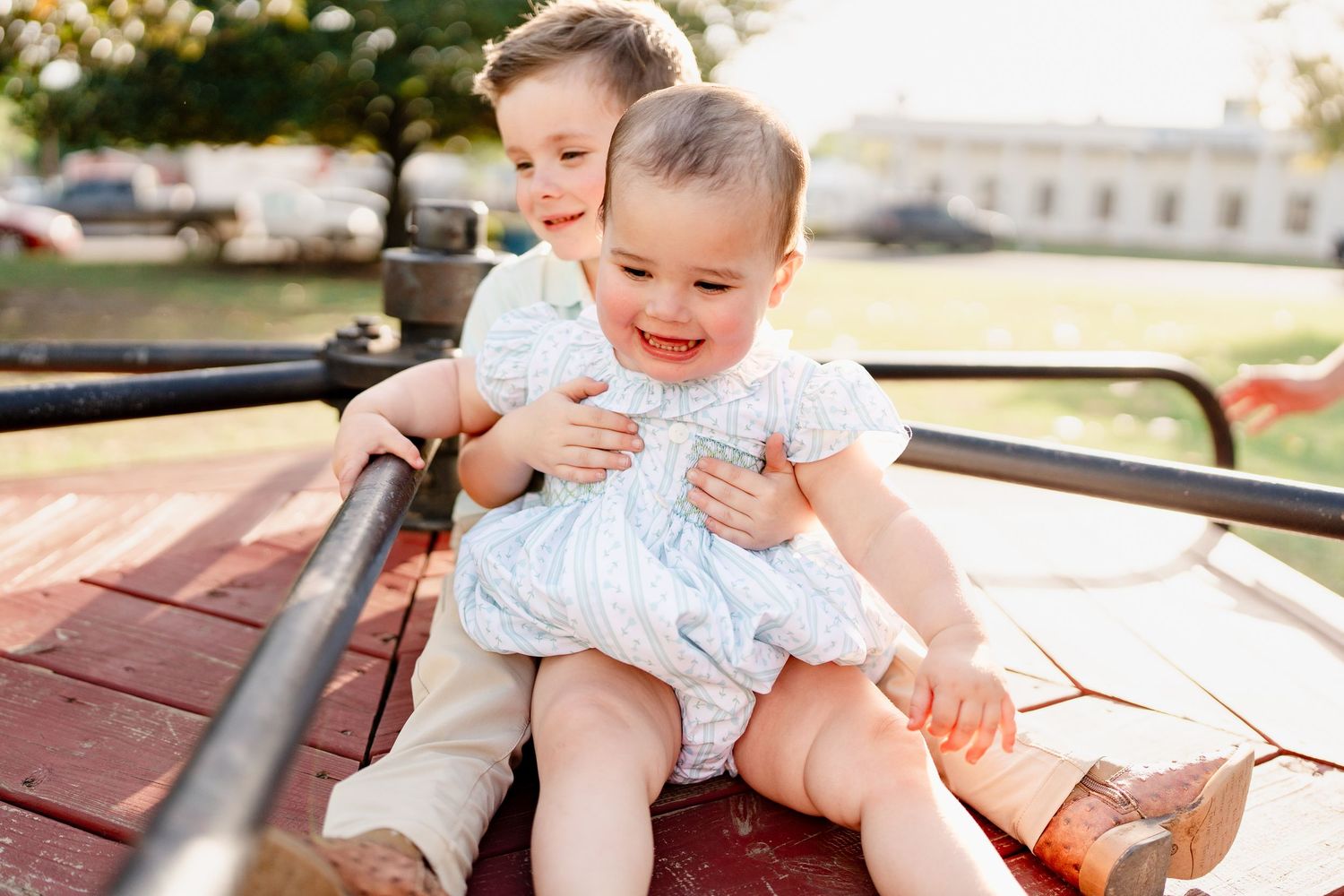 Two children sit together on a red surface outdoors during a sunny day.