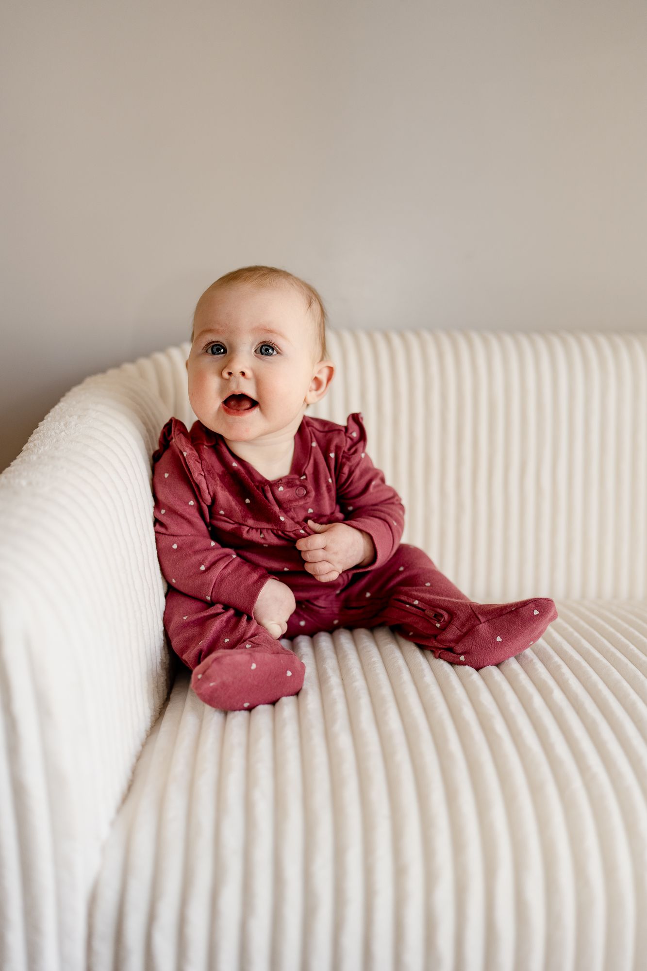 Baby in burgundy outfit lying playfully on white textured couch.