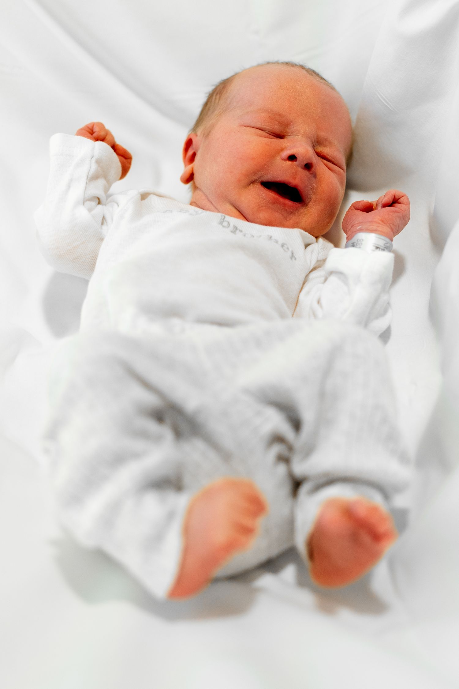 Newborn baby wearing white outfit lying on white bedding while stretching and yawning.