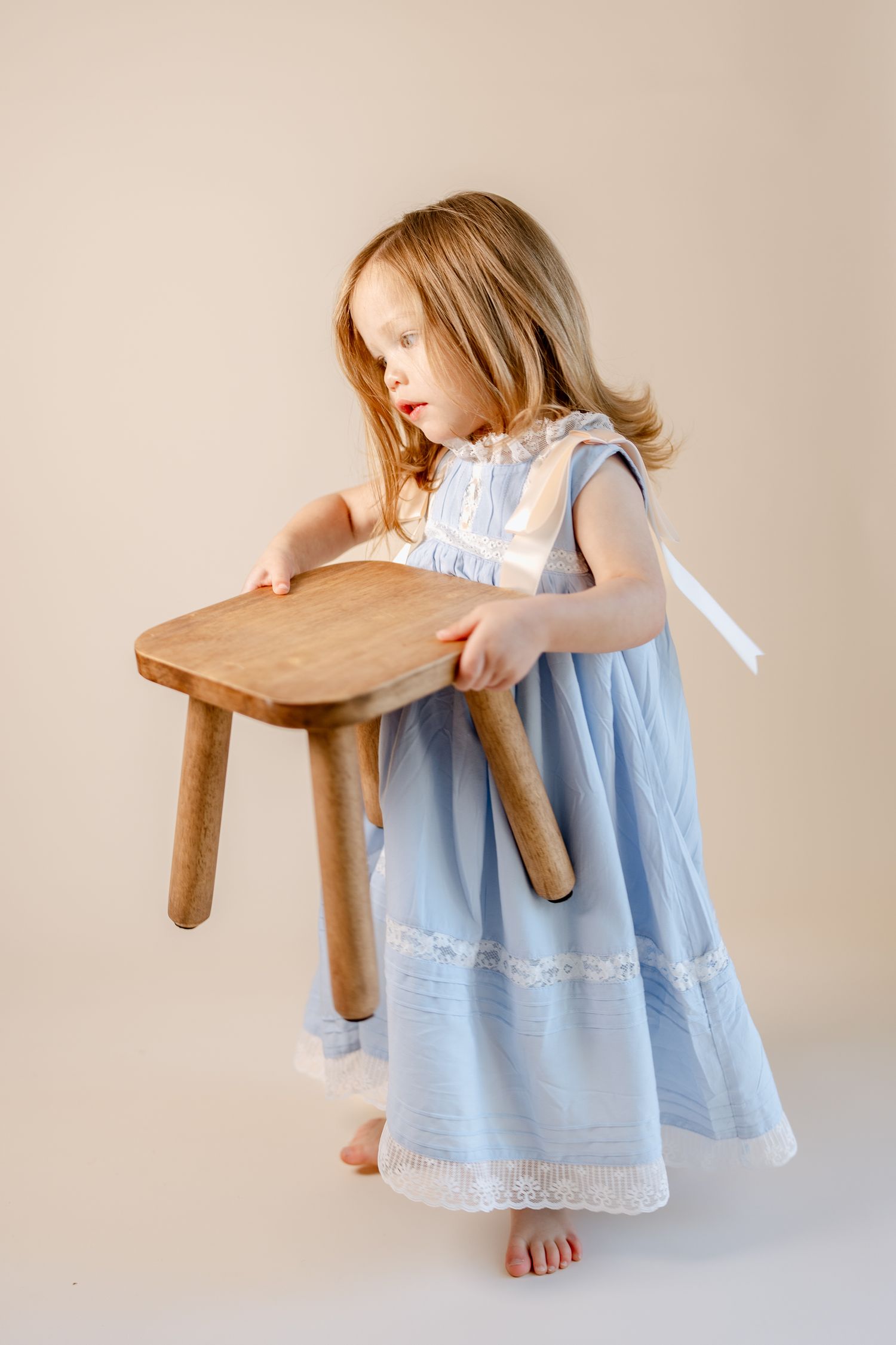 Side view of child holding wooden stool while wearing blue lace-trimmed dress.