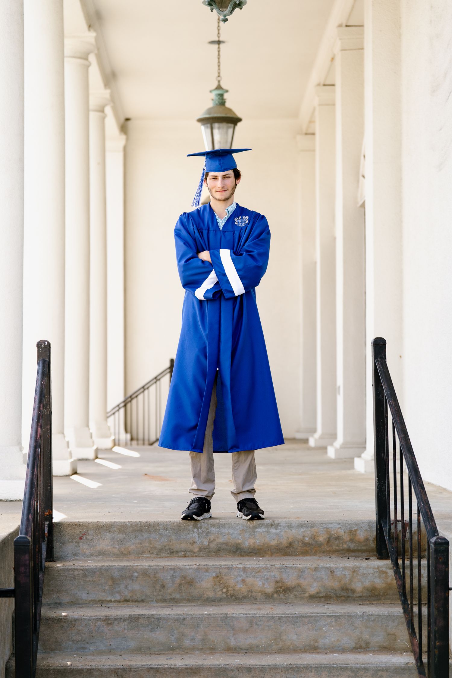 Student in graduation attire poses between white columns with arms crossed.