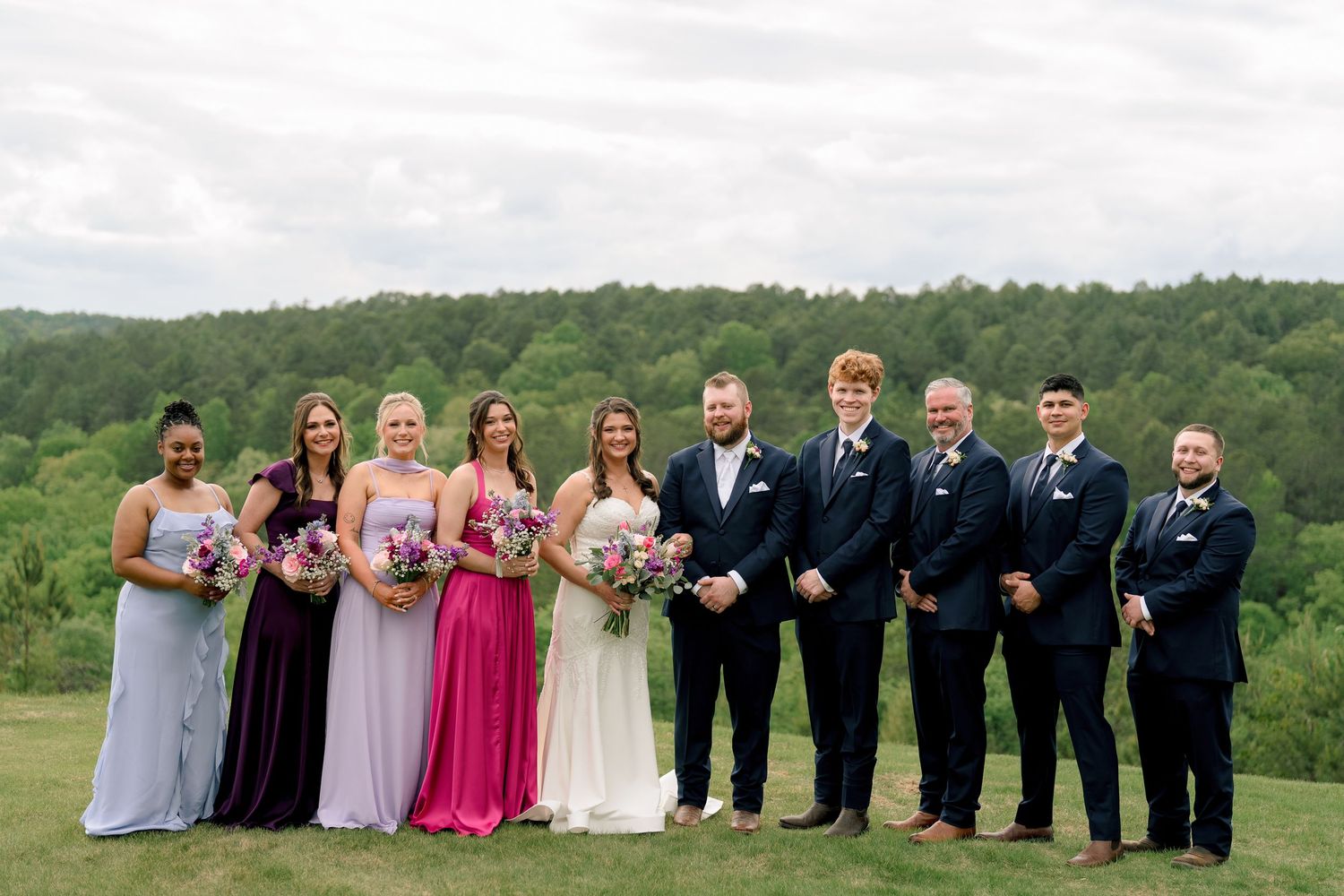 Wedding party poses outdoors with bridesmaids in purple dresses and groomsmen in navy suits against green hills.