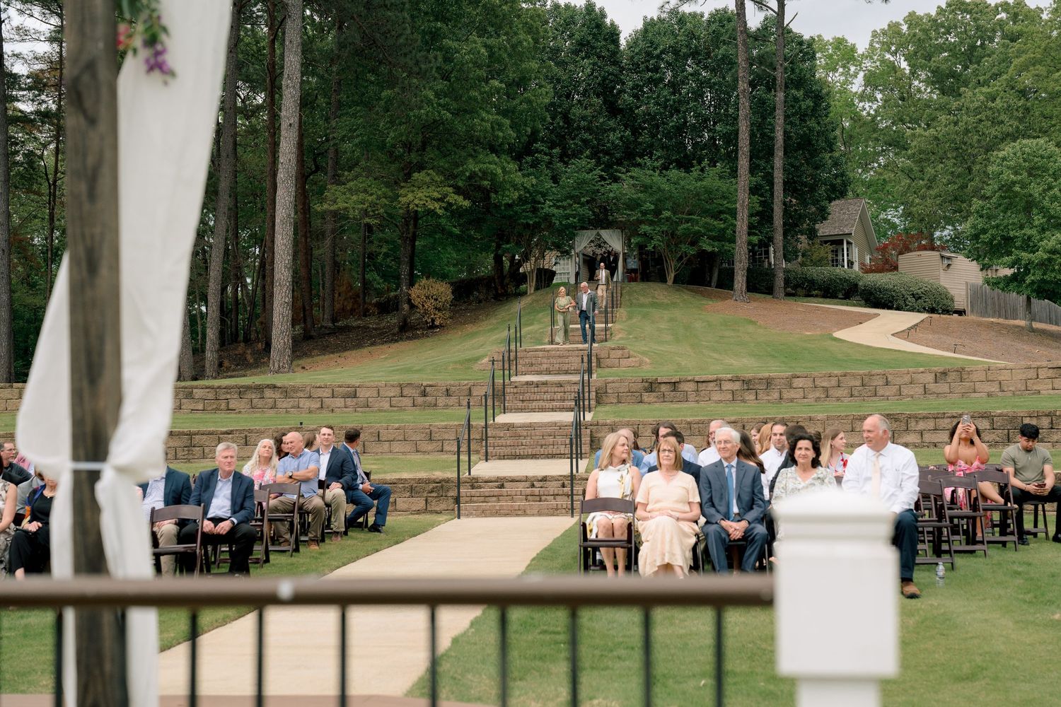Outdoor wedding ceremony with guests seated on chairs along an aisle, surrounded by lush green trees and terraced steps.