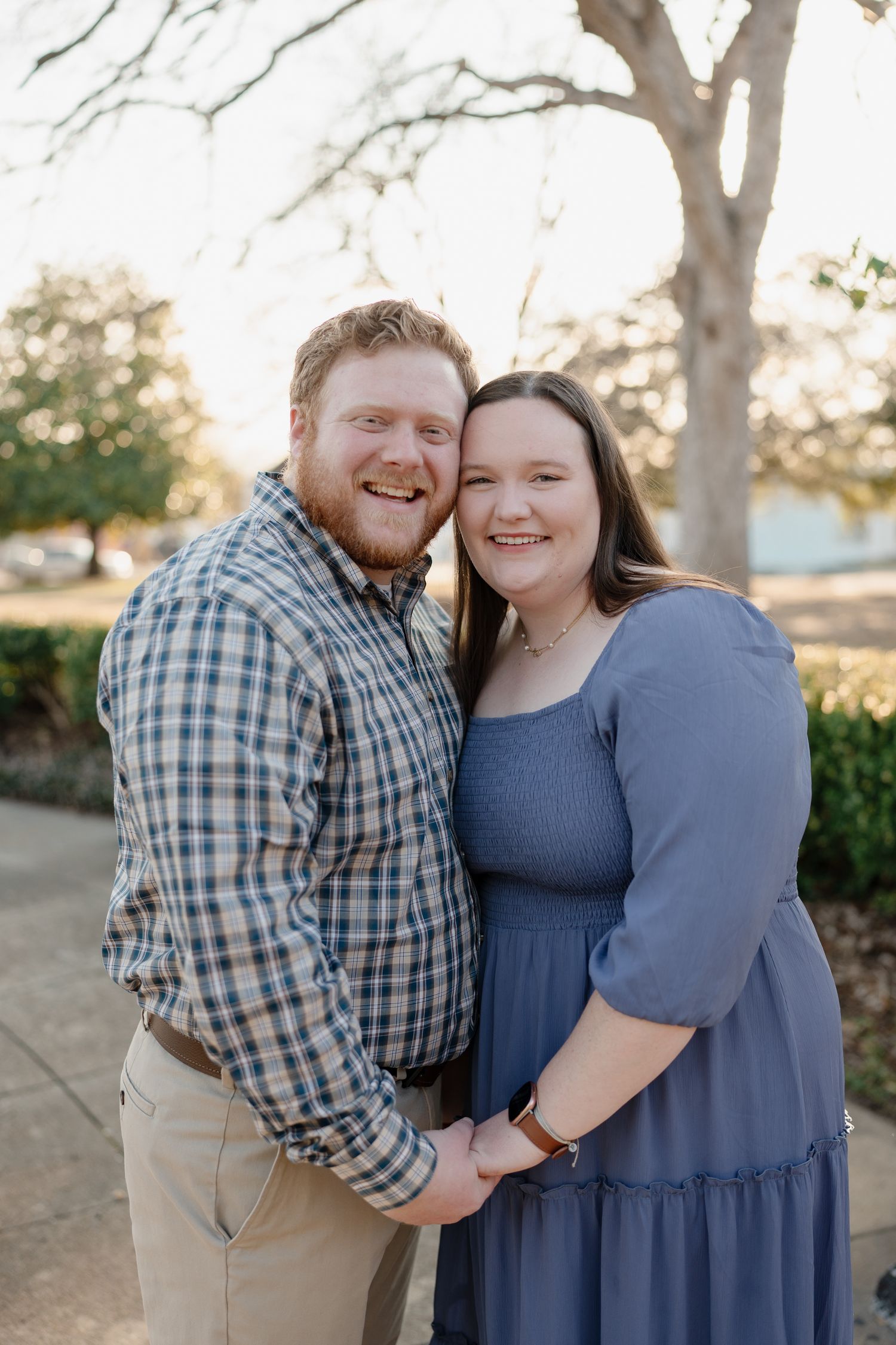 Smiling couple posing outdoors, man in plaid shirt and woman in blue dress, holding hands in a sunny park setting.