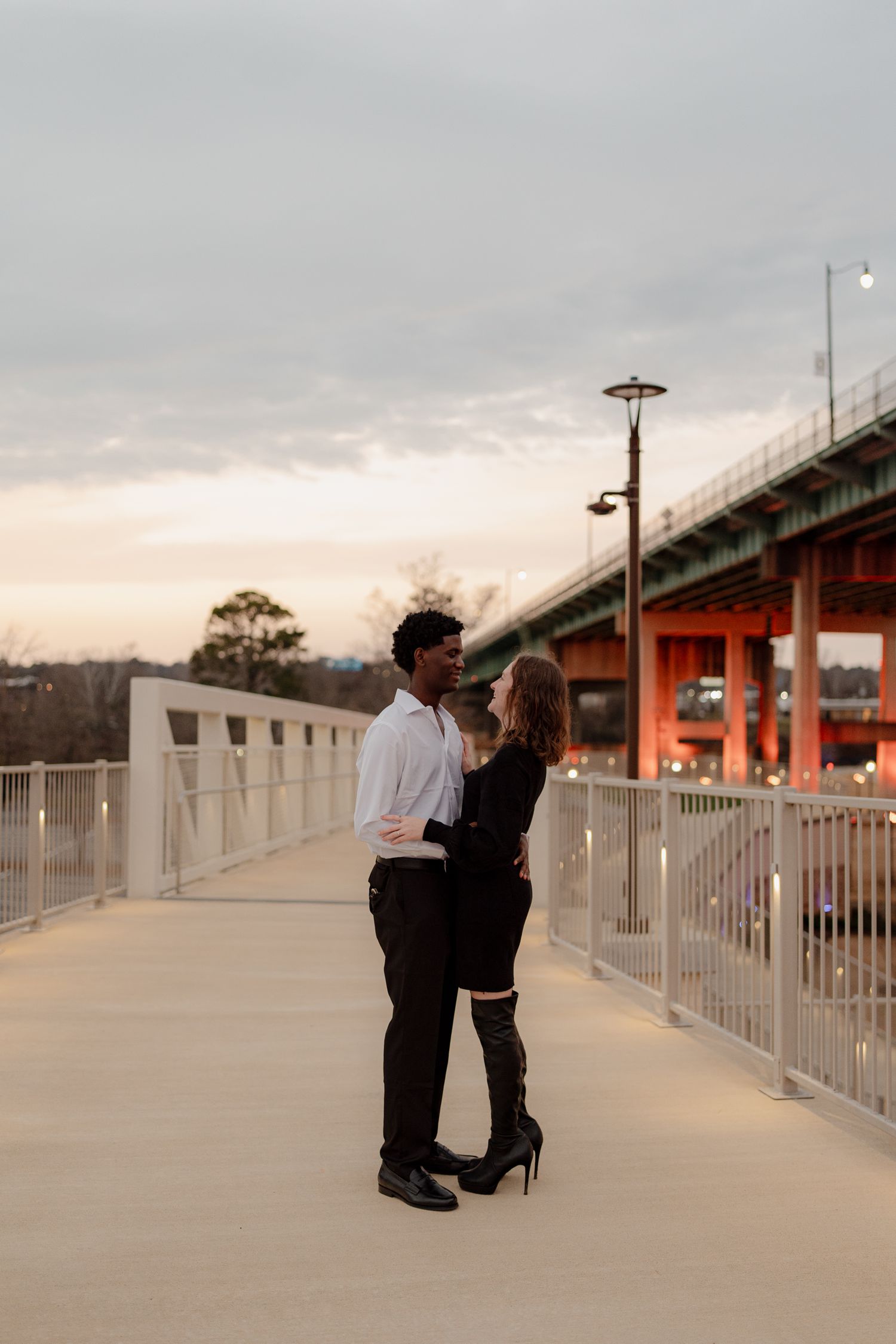 Romantic interracial couple embracing on a pedestrian bridge at sunset, woman in heels and man in white shirt and black pants.