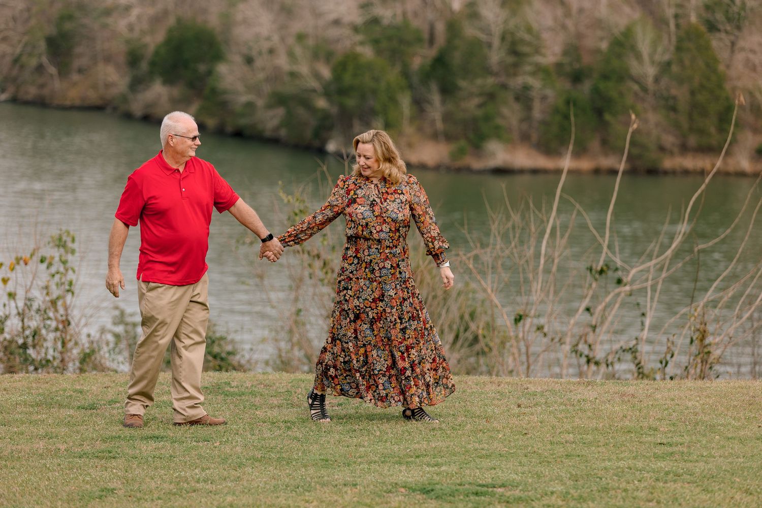 Older couple holding hands and walking together on a grassy lawn beside a peaceful river.