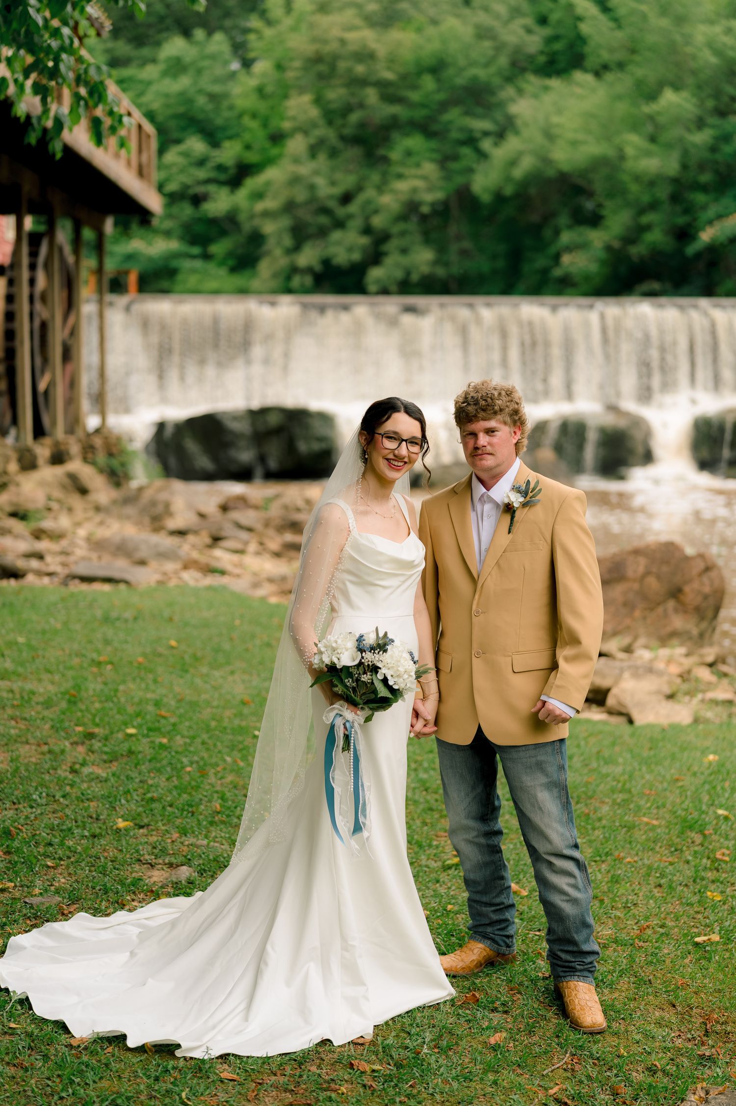 Bride and groom pose together in front of scenic waterfall, bride holding white and blue bouquet.