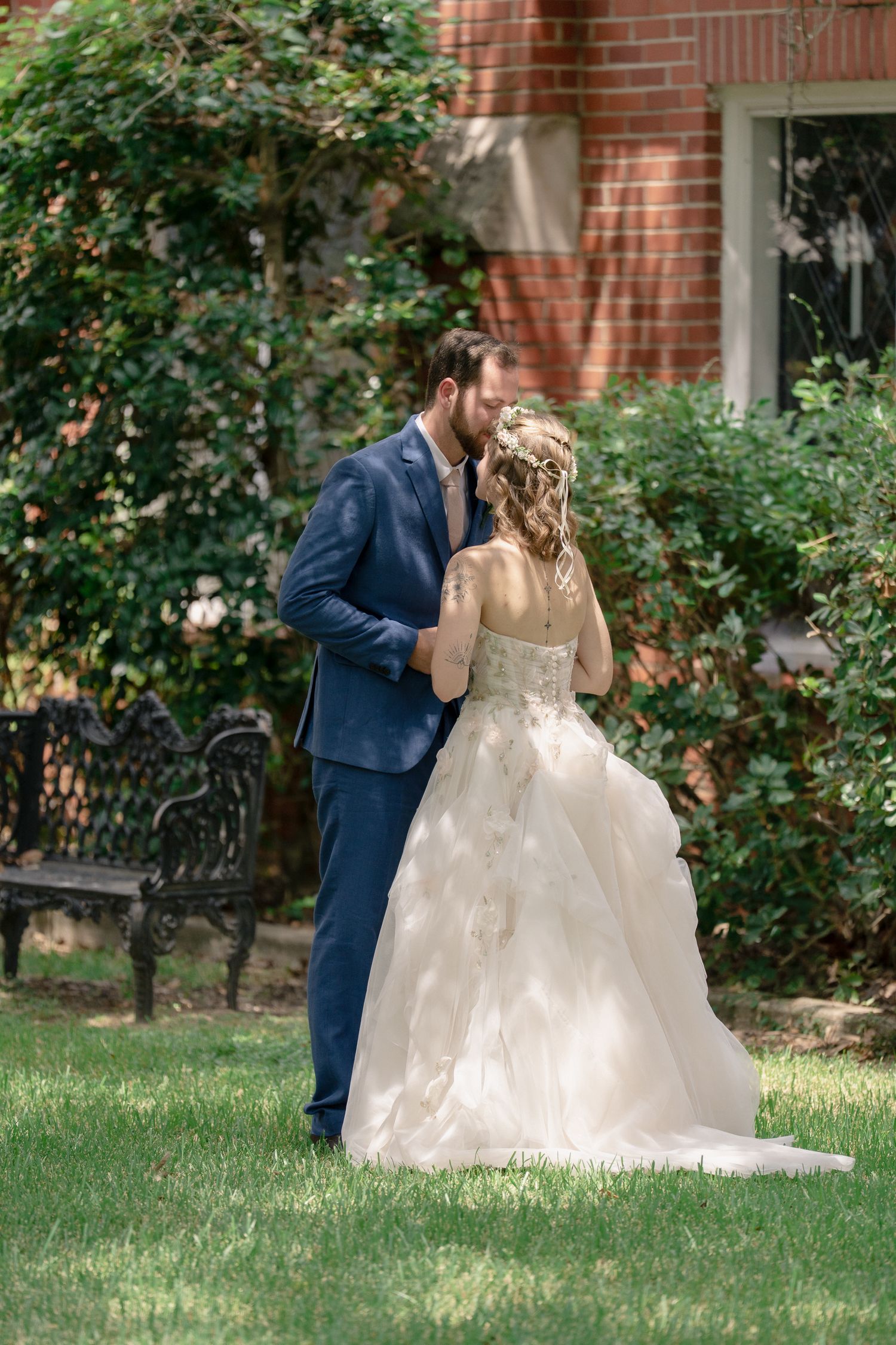 A couple in wedding attire shares a tender moment in a garden setting with brick buildings and greenery.