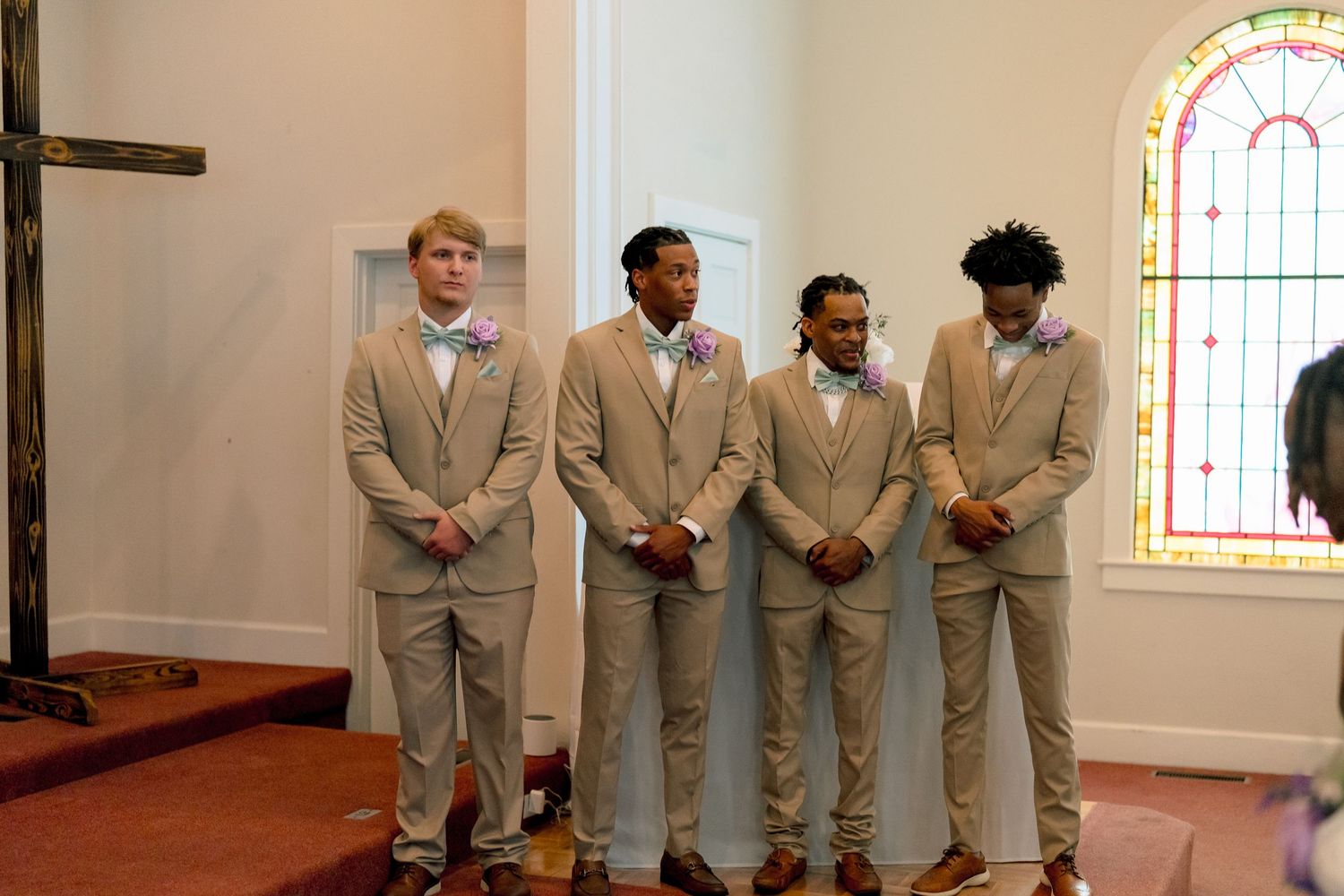 Four groomsmen in matching tan suits with colorful bow ties standing at church altar before wedding ceremony.