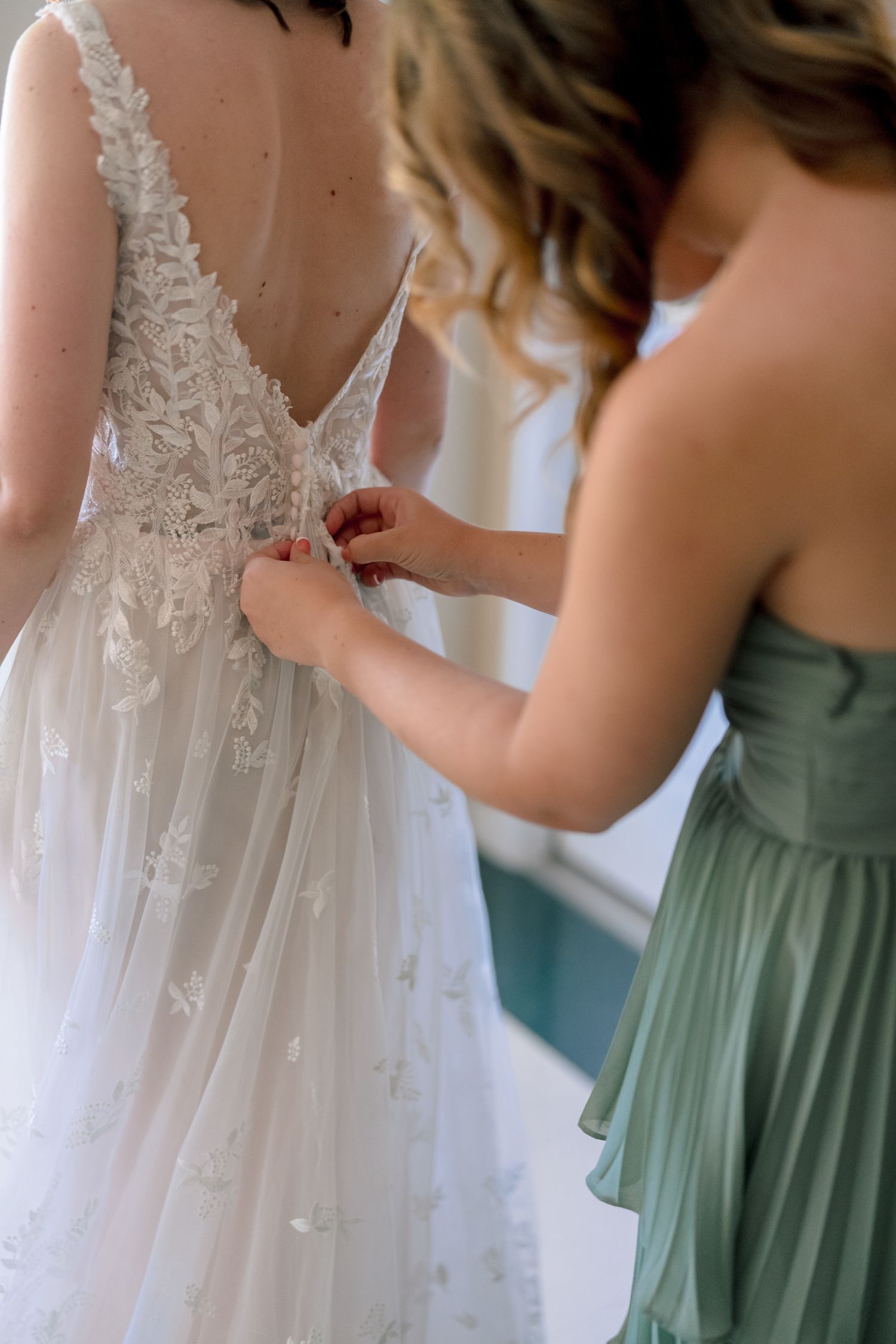 Bridesmaid in green dress fastening the back of a white lace wedding gown on the bride.